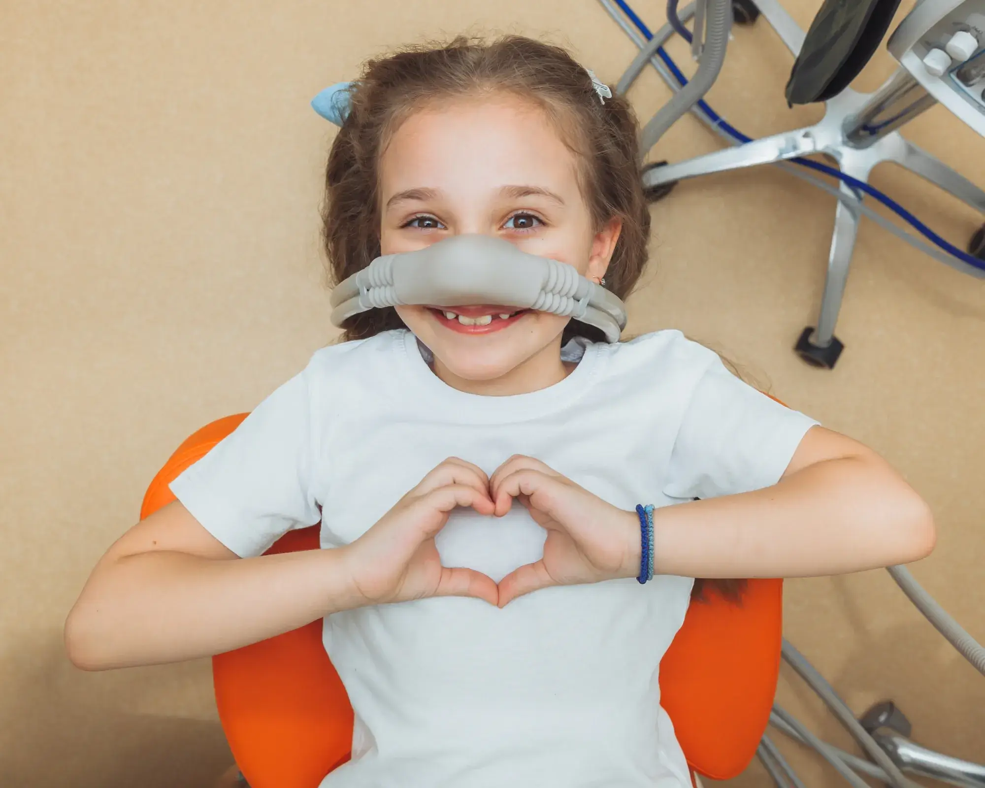 Young girl at dentist with a laughing gas mask, smiling and forming a heart shape with hands.
