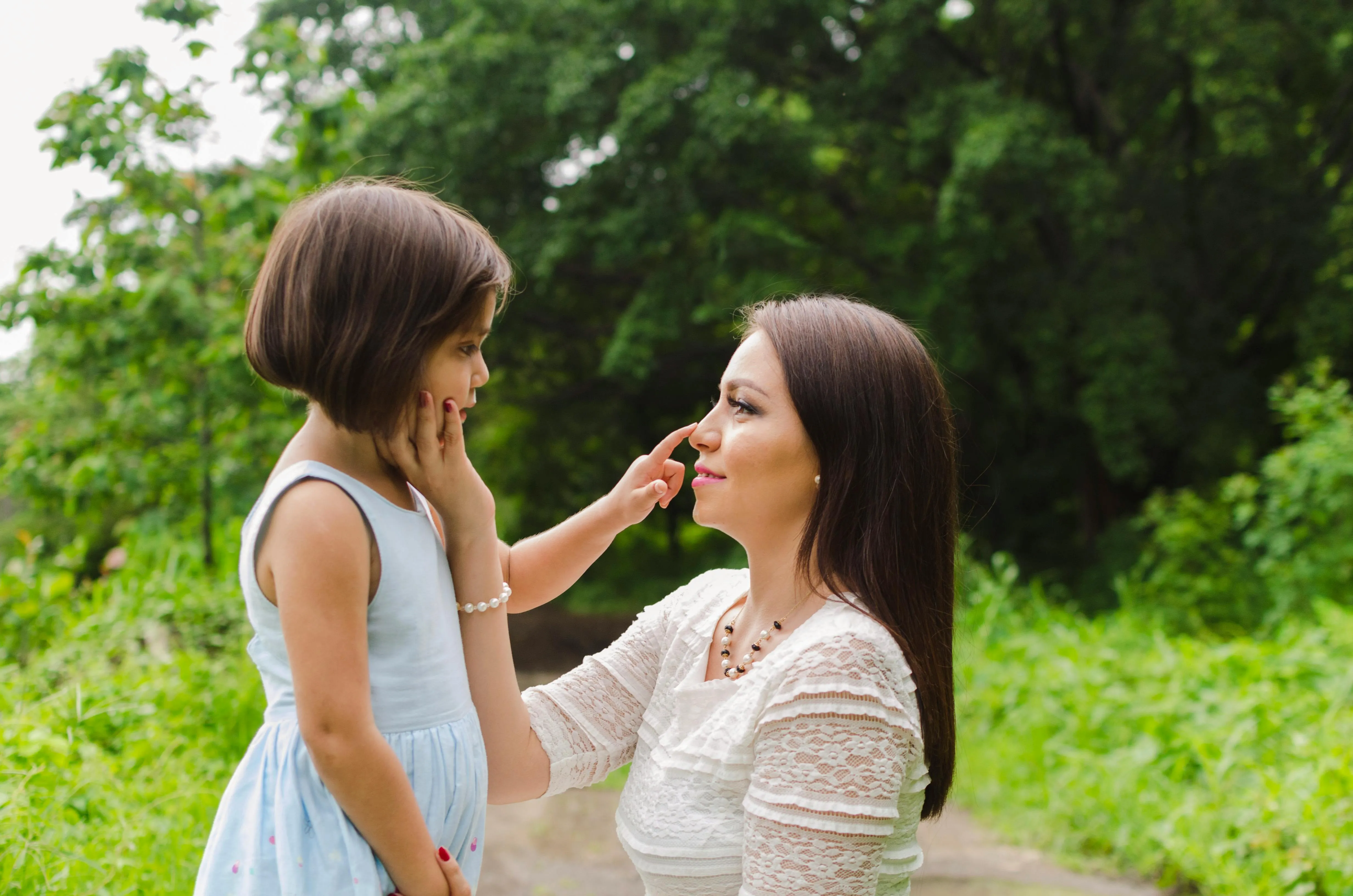Young girl in a blue dress touching the nose of a woman in a white lace top, outdoors with green foliage in the background.