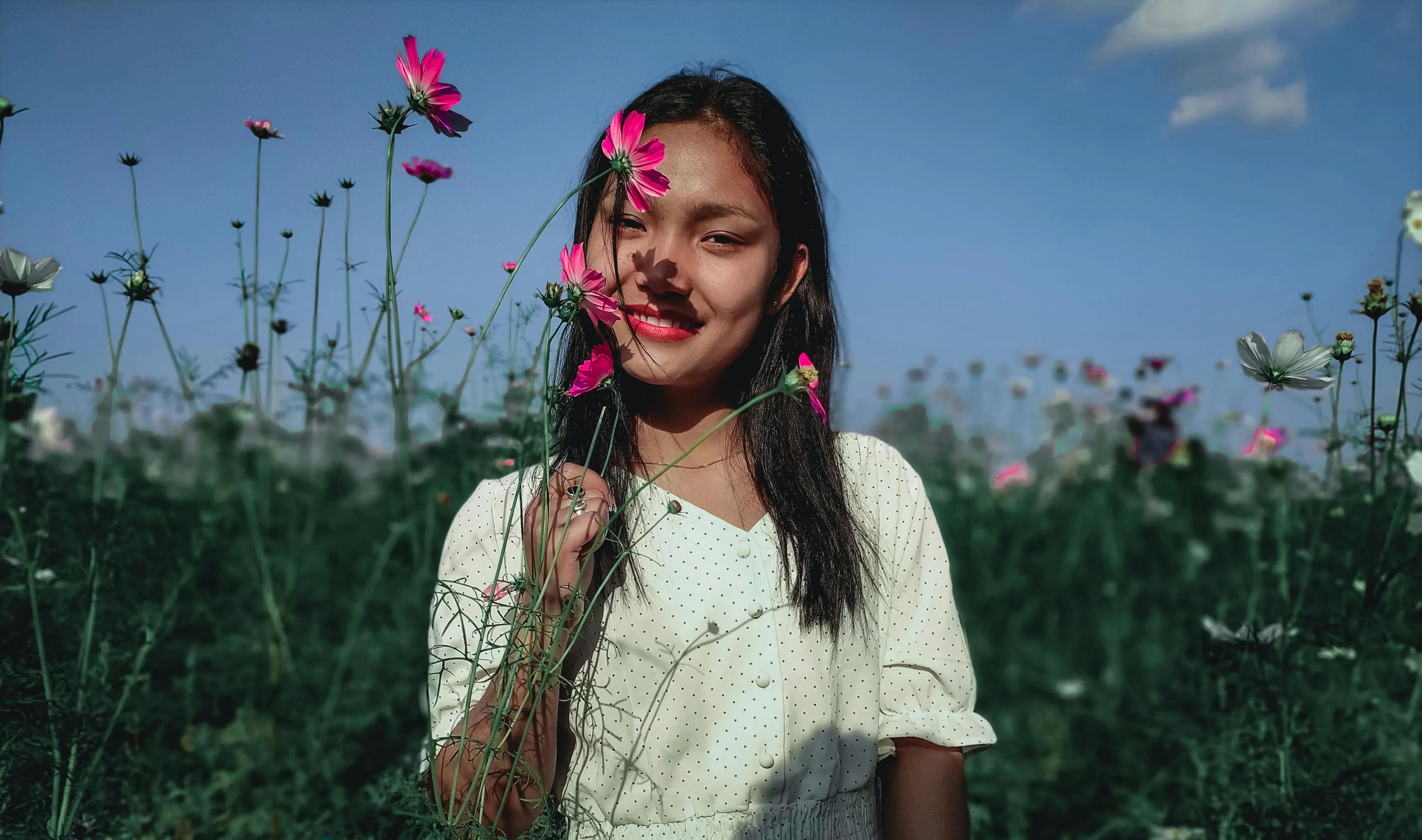 Smiling woman with long black hair holding pink flowers in a green field under a blue sky.