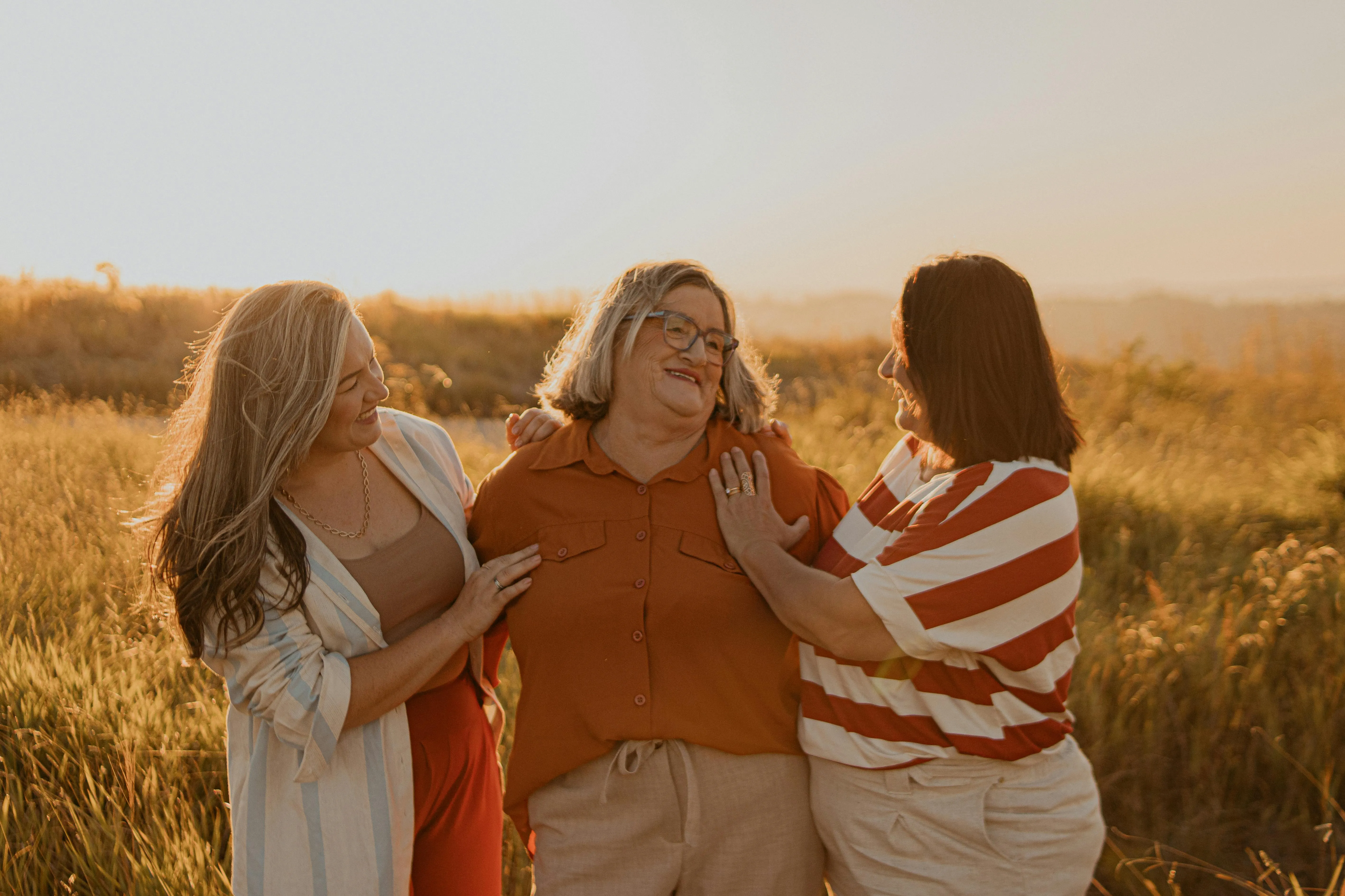 Three women smiling and standing closely together in a sunlit grassy field during golden hour.