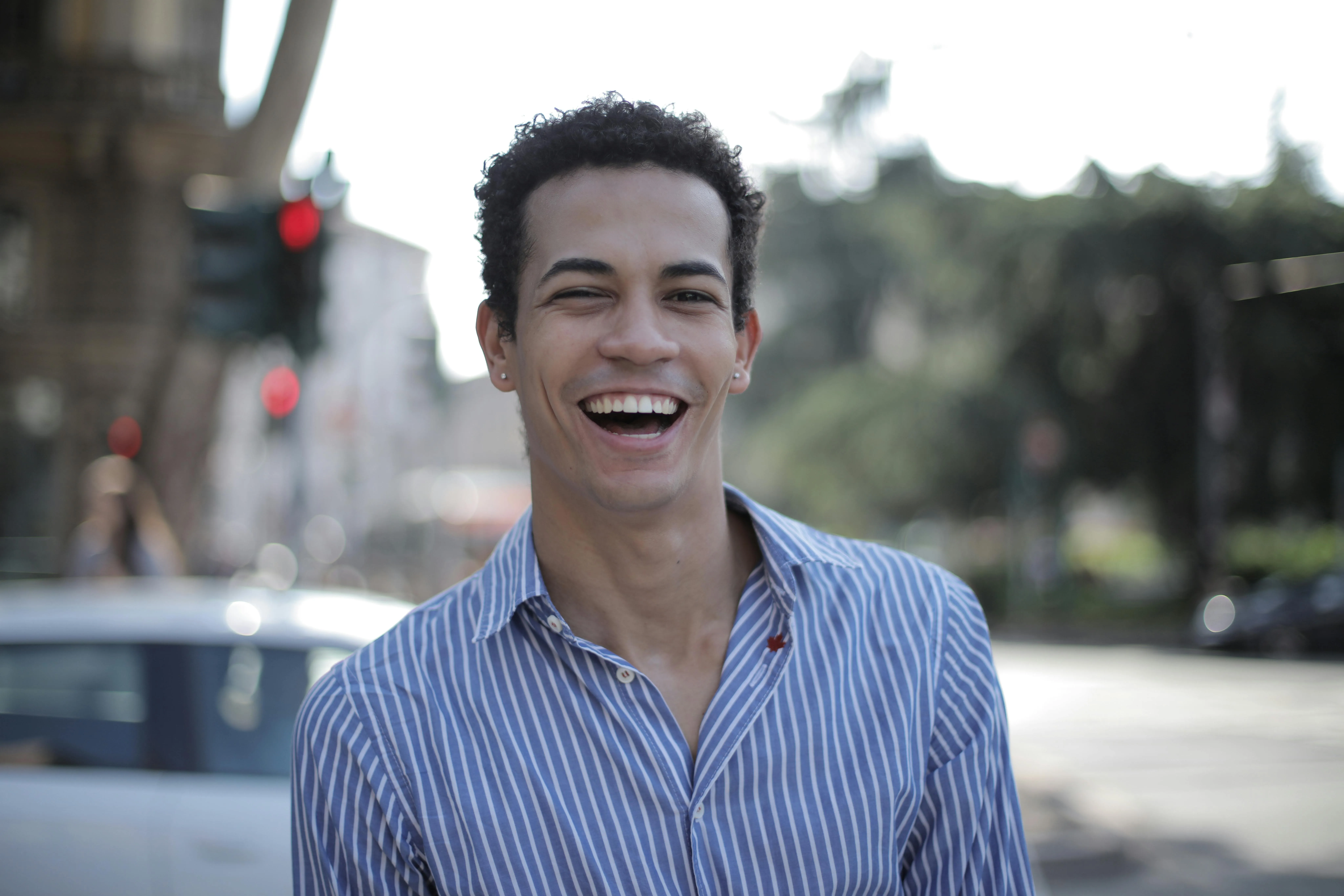Smiling young man in a blue and white striped shirt standing outdoors with blurred city background.