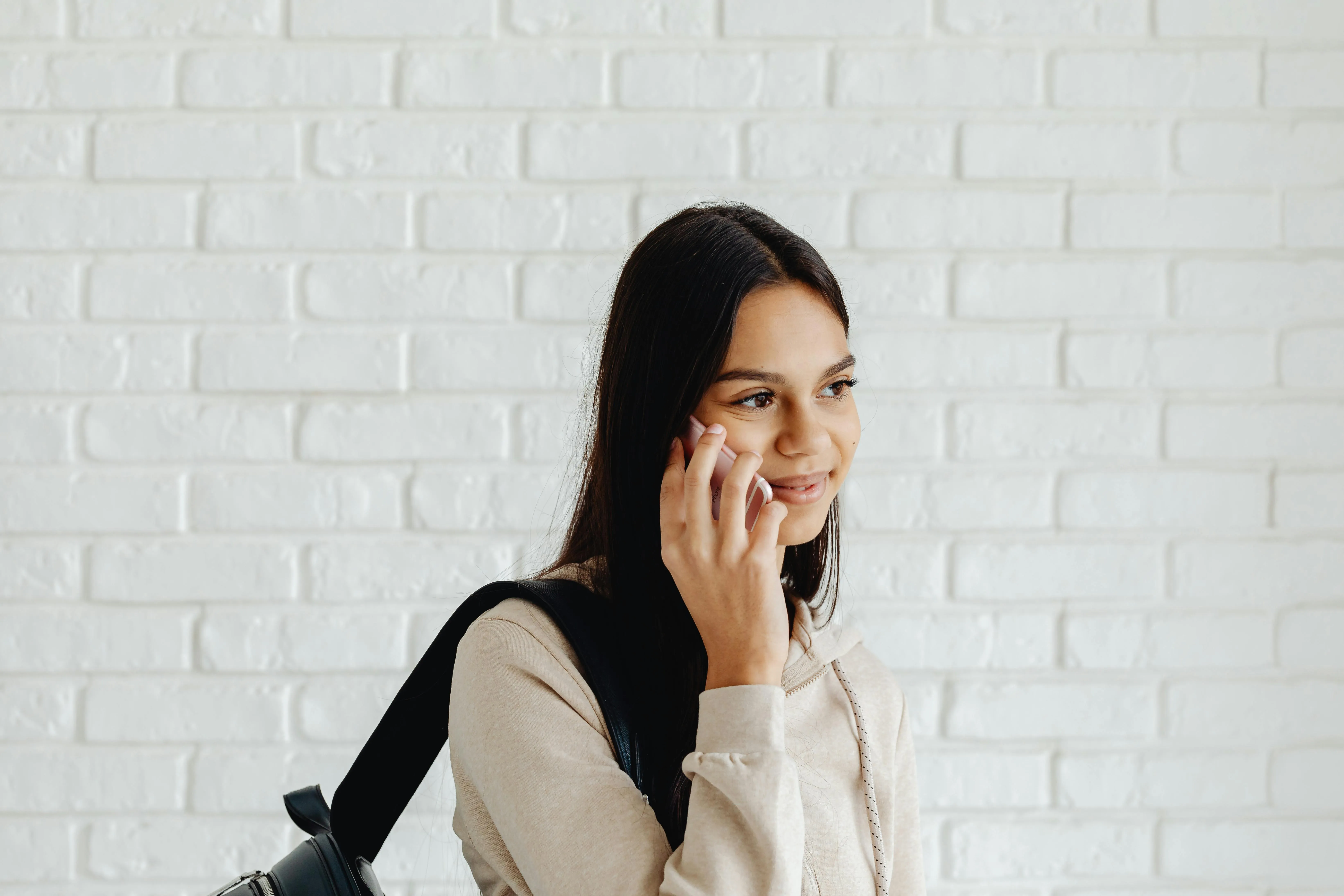 Young woman with long dark hair, wearing a beige hoodie and black backpack, smiling while talking on a smartphone against a white brick wall.