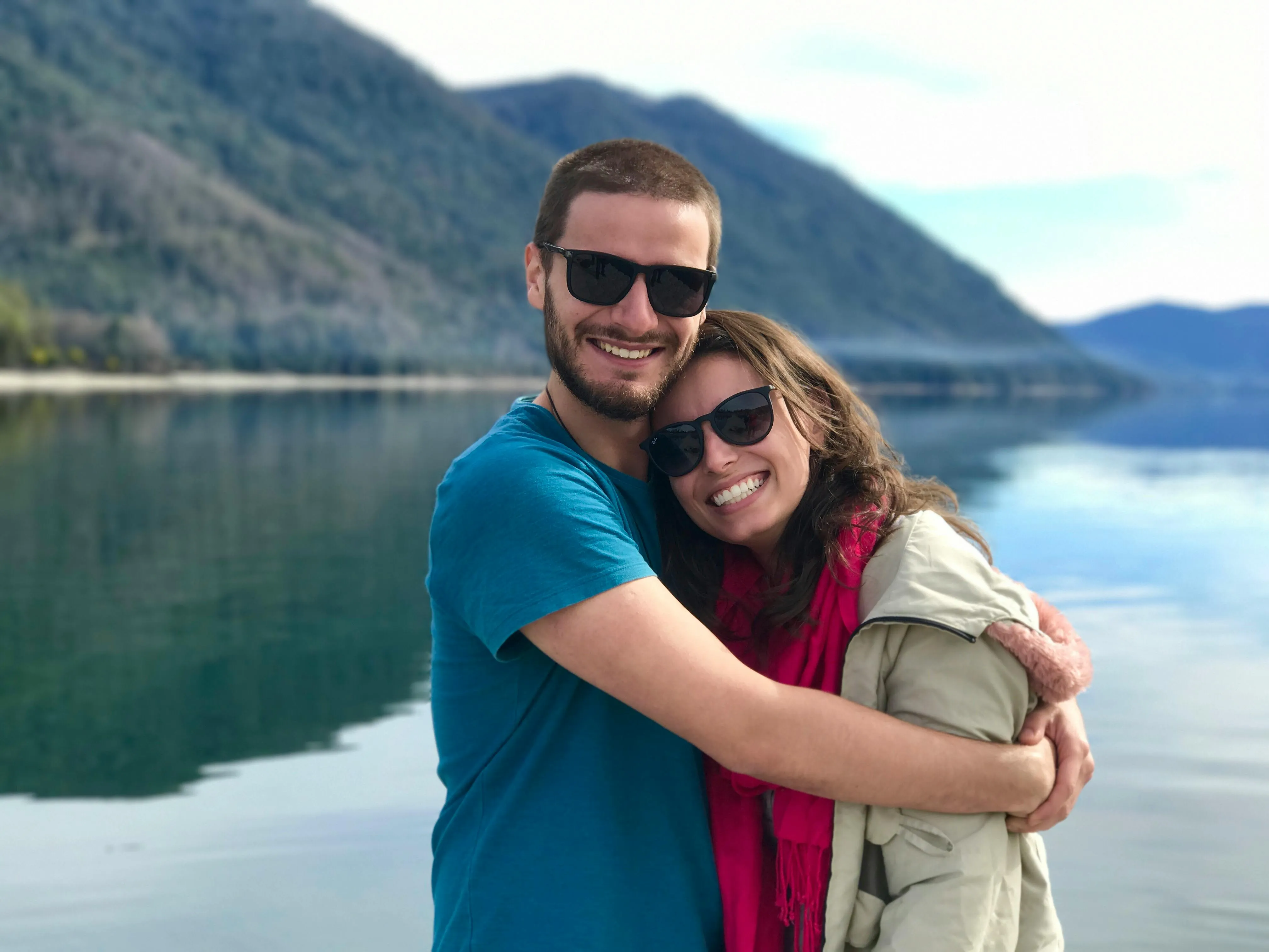 Smiling couple wearing sunglasses hugging near a calm lake with mountains in the background.