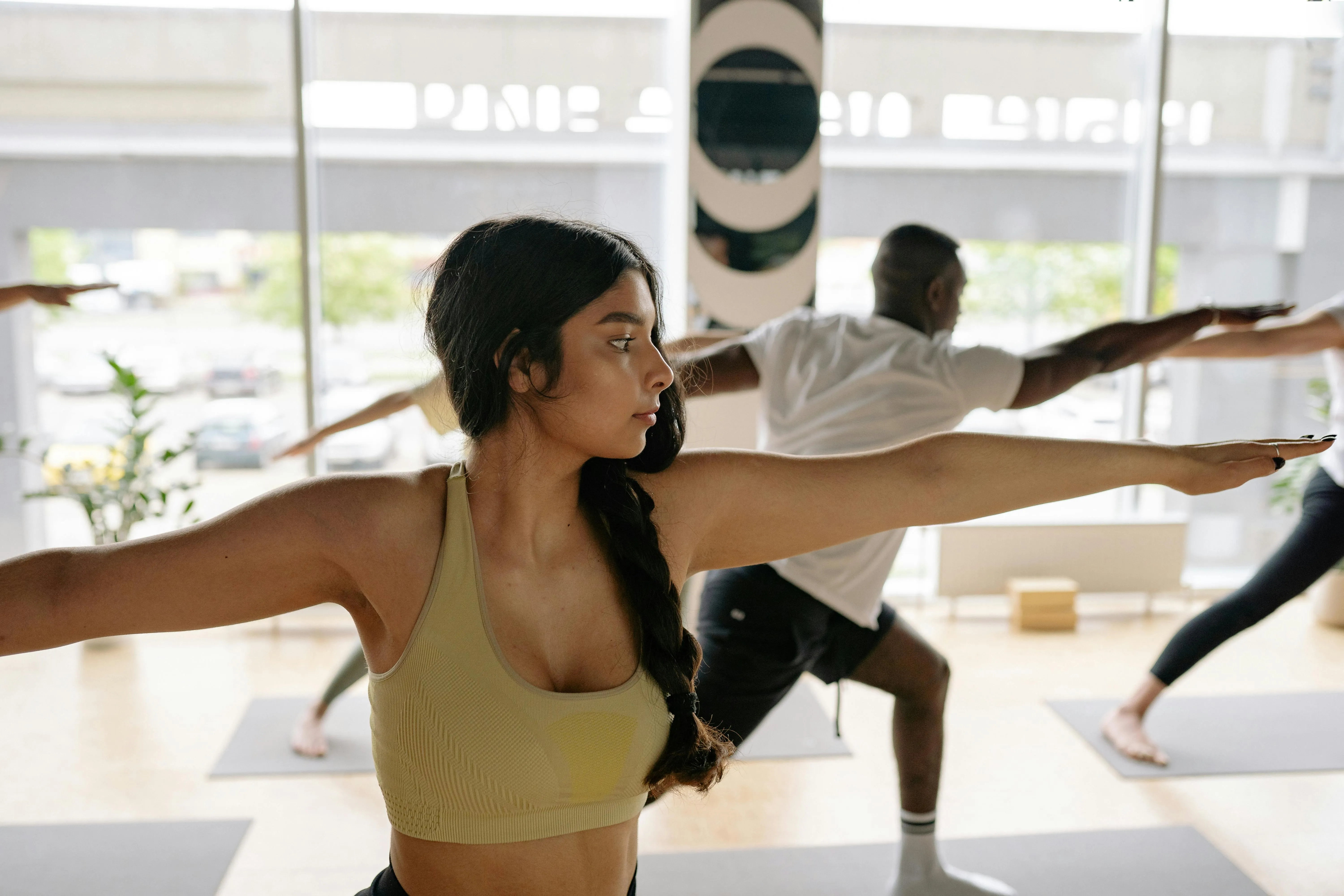A woman with a braid wearing a yellow sports bra practices yoga in warrior pose in a bright studio with others.