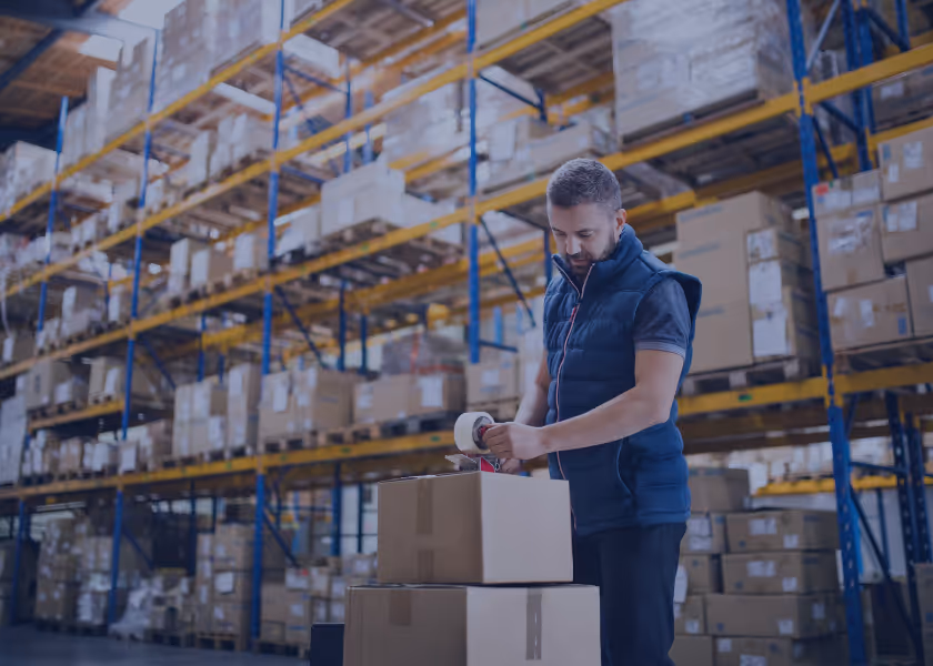 Mann in Weste klebt Verpackungsklebeband auf Pappkartons in einem Lager mit hohen Regalen voller Kartons.