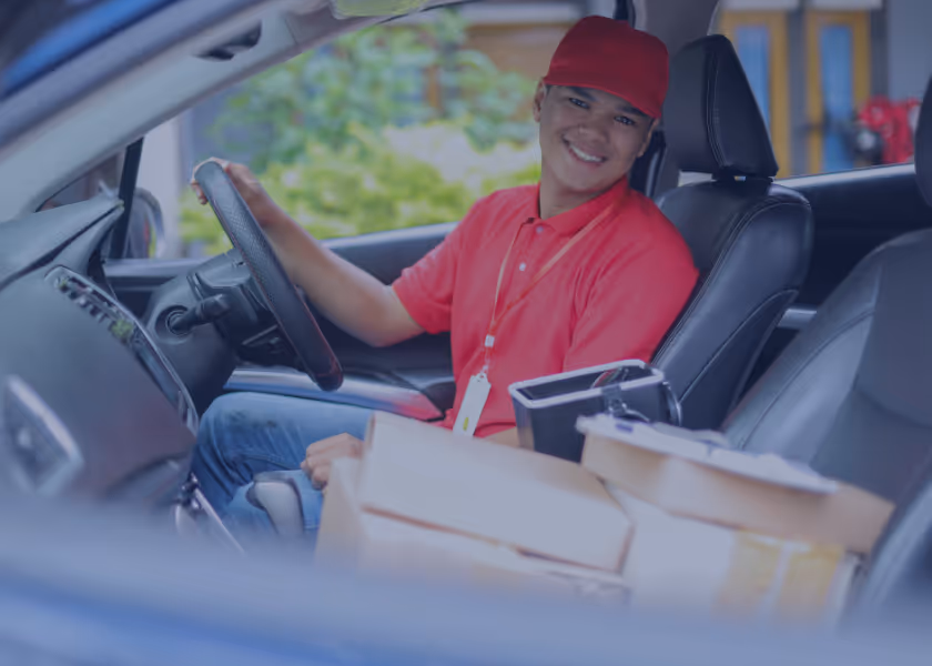 Ein Lieferfahrer in rotem Hemd und roter Mütze sitzt lächelnd im Fahrzeug hinter dem Steuer, vor ihm liegen mehrere Pakete.