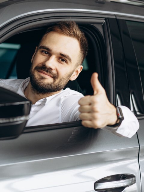 Șofer cu barbă la volanul unei mașini gri, cu degetul mare ridicat