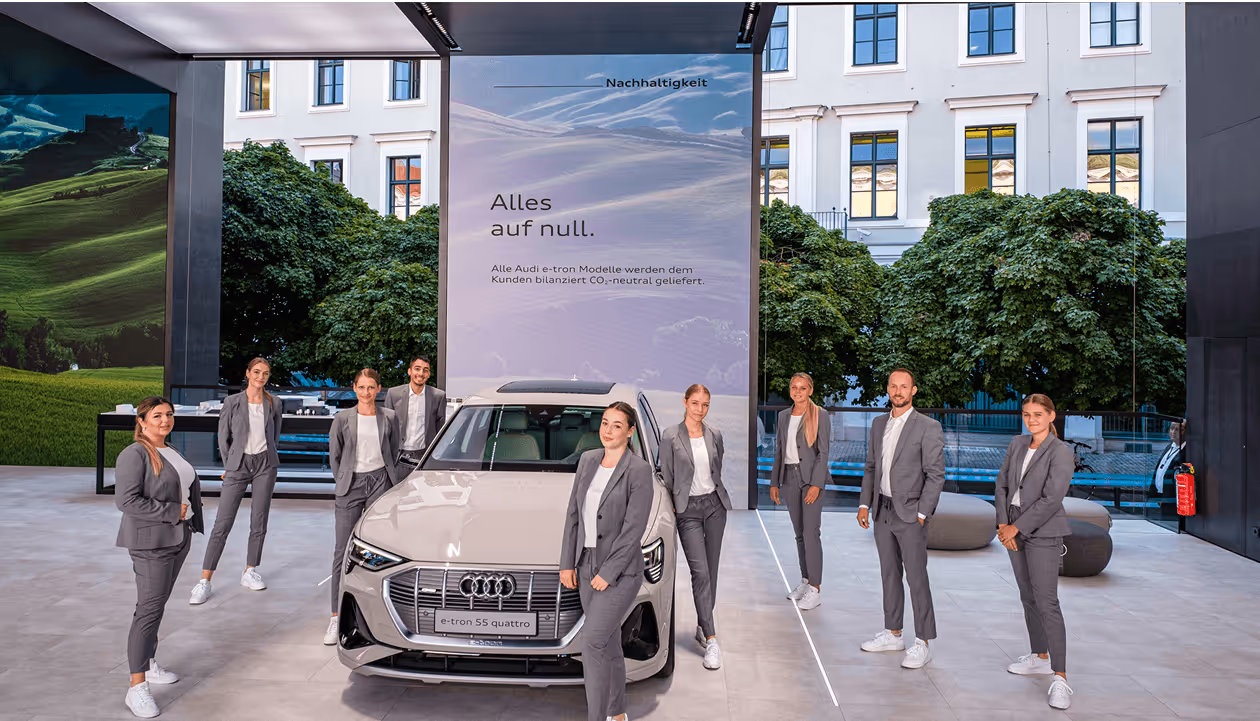 Personengruppe in grauen Anzügen vor einem Elektroauto in einem modernen Showroom mit großem Bildschirm und nachhaltiger Botschaft.