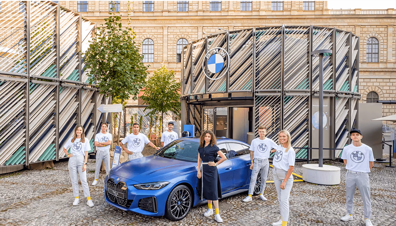 Blaues Auto vor modernem Gebäude, umgeben von acht lächelnden Personen in weißer Kleidung.