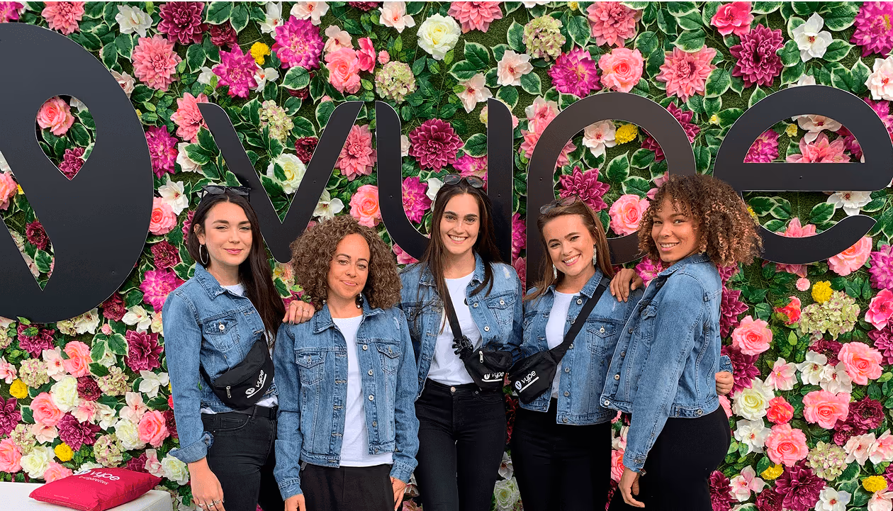 Fünf Frauen in Jeansjacken vor bunter Blumenwand mit großem Schriftzug.