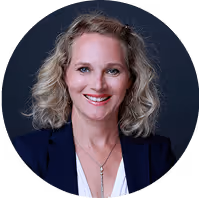 Blonde Frau mit welligem Haar und Blazer lächelt vor dunklem Hintergrund.