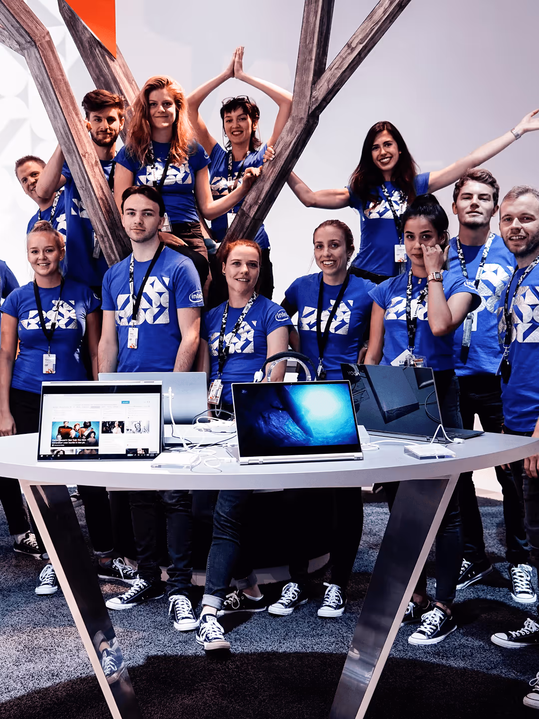 Gruppe junger Erwachsener in blauen T-Shirts posiert fröhlich um einen Tisch mit Laptops auf einem Messe-Stand.