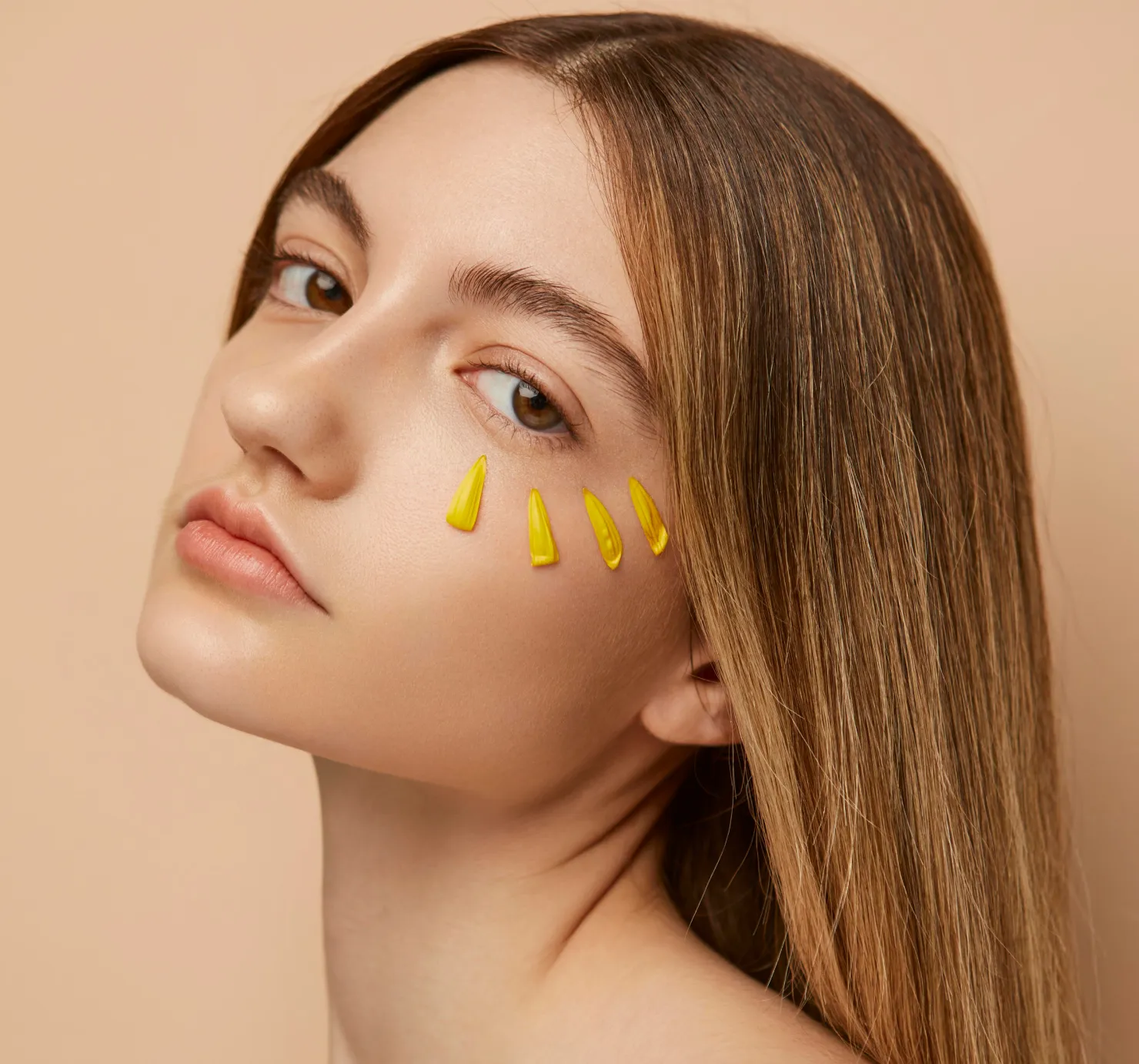 Close-up portrait of a young woman with long brown hair, looking at the camera, with five small yellow, sun ray-like dollops of skincare product applied under her eye.