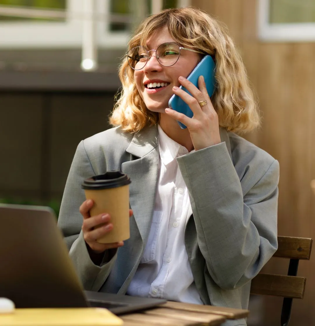 A smiling woman with blonde, wavy hair and glasses is holding a coffee cup and talking on a blue smartphone while sitting outdoors near a laptop.