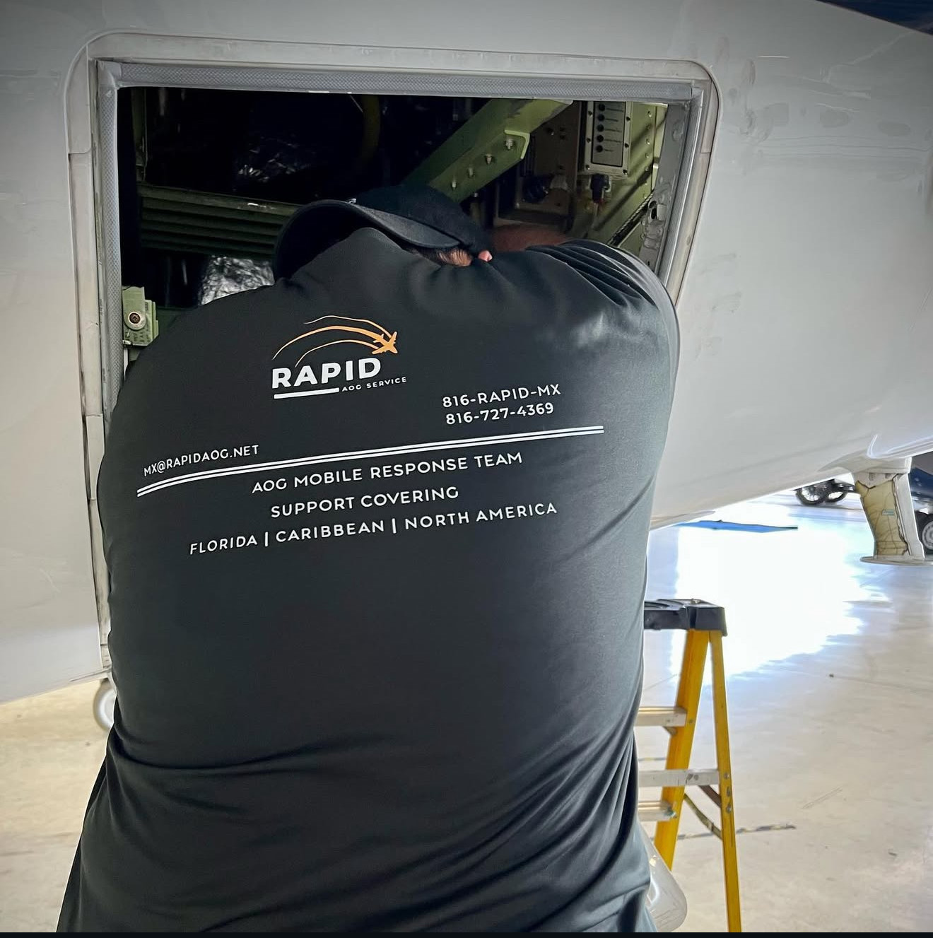 Person wearing a black Rapid AOG Service shirt working on an aircraft panel inside a hangar.