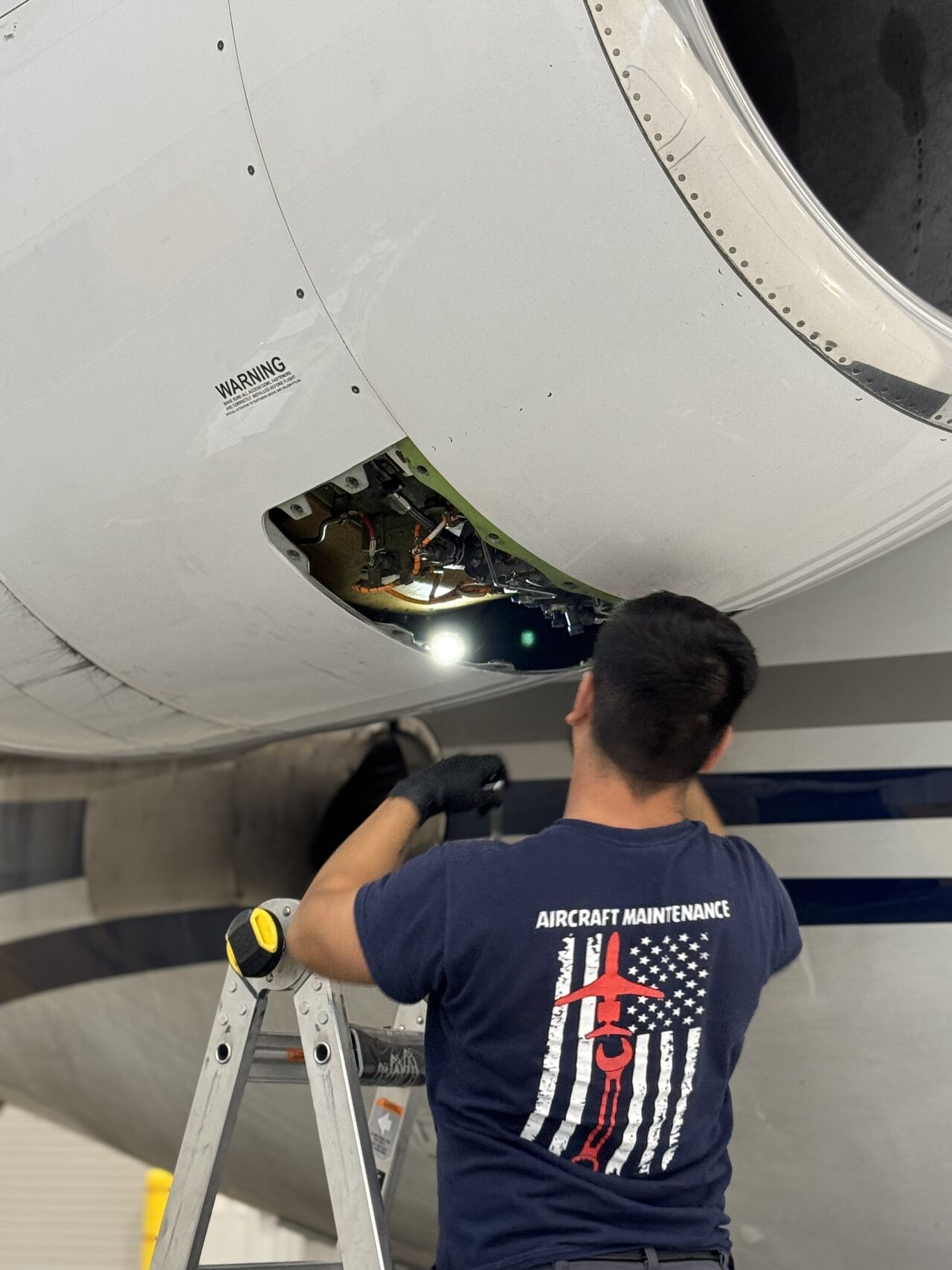 Aircraft maintenance worker on a ladder inspecting an open panel on an airplane engine.