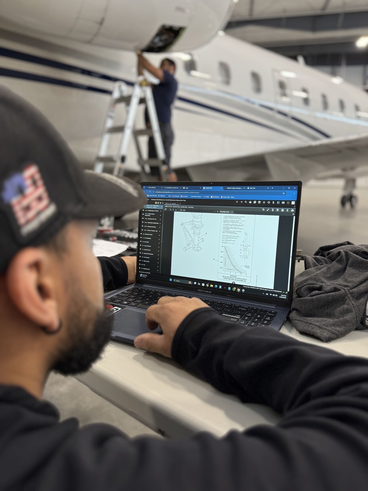 Man wearing a cap works on a laptop displaying aircraft maintenance diagrams inside an airplane hangar with another person on a ladder inspecting a plane engine.
