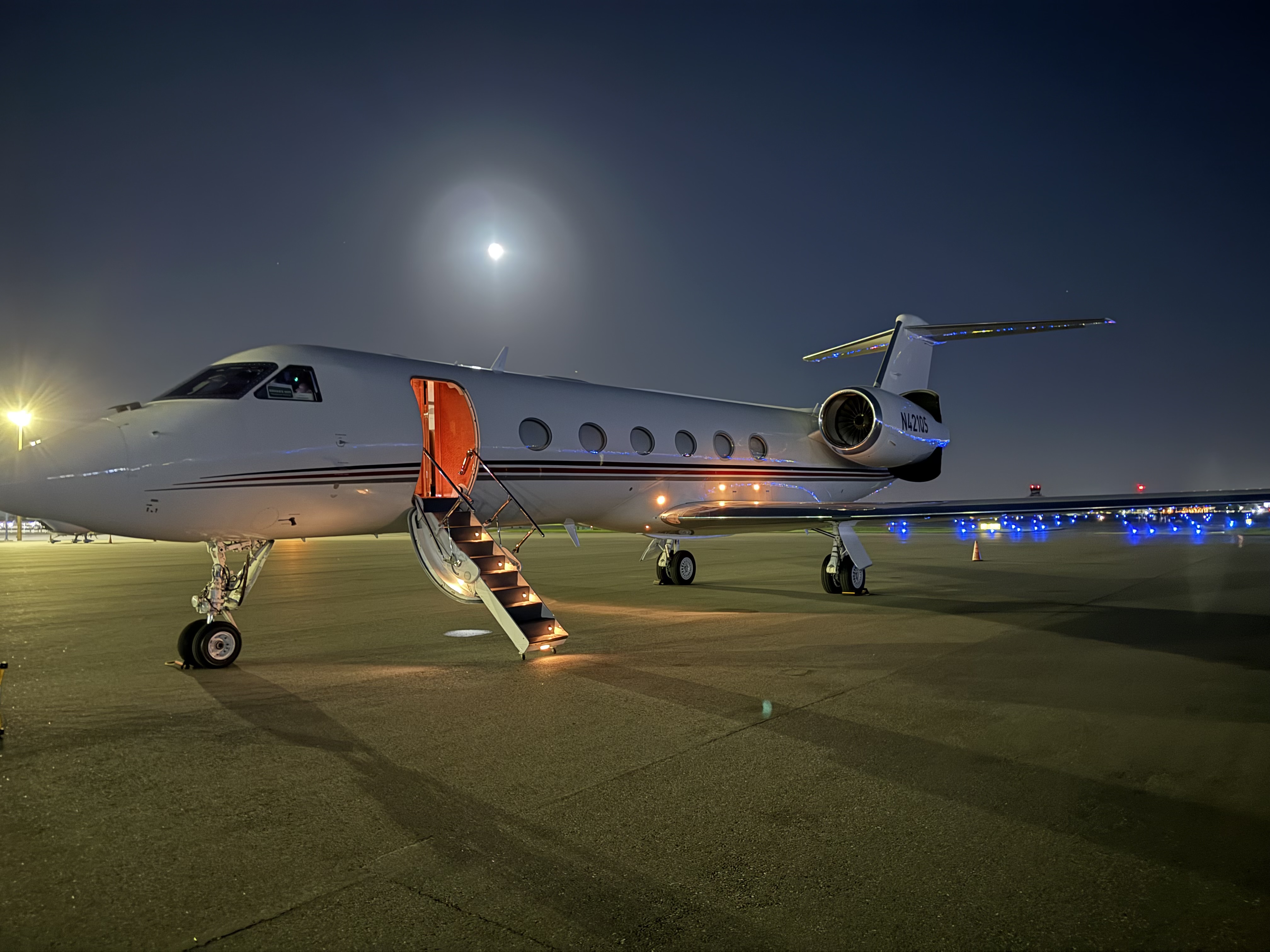Private jet with open illuminated stairs parked on airport tarmac under a full moon at night.