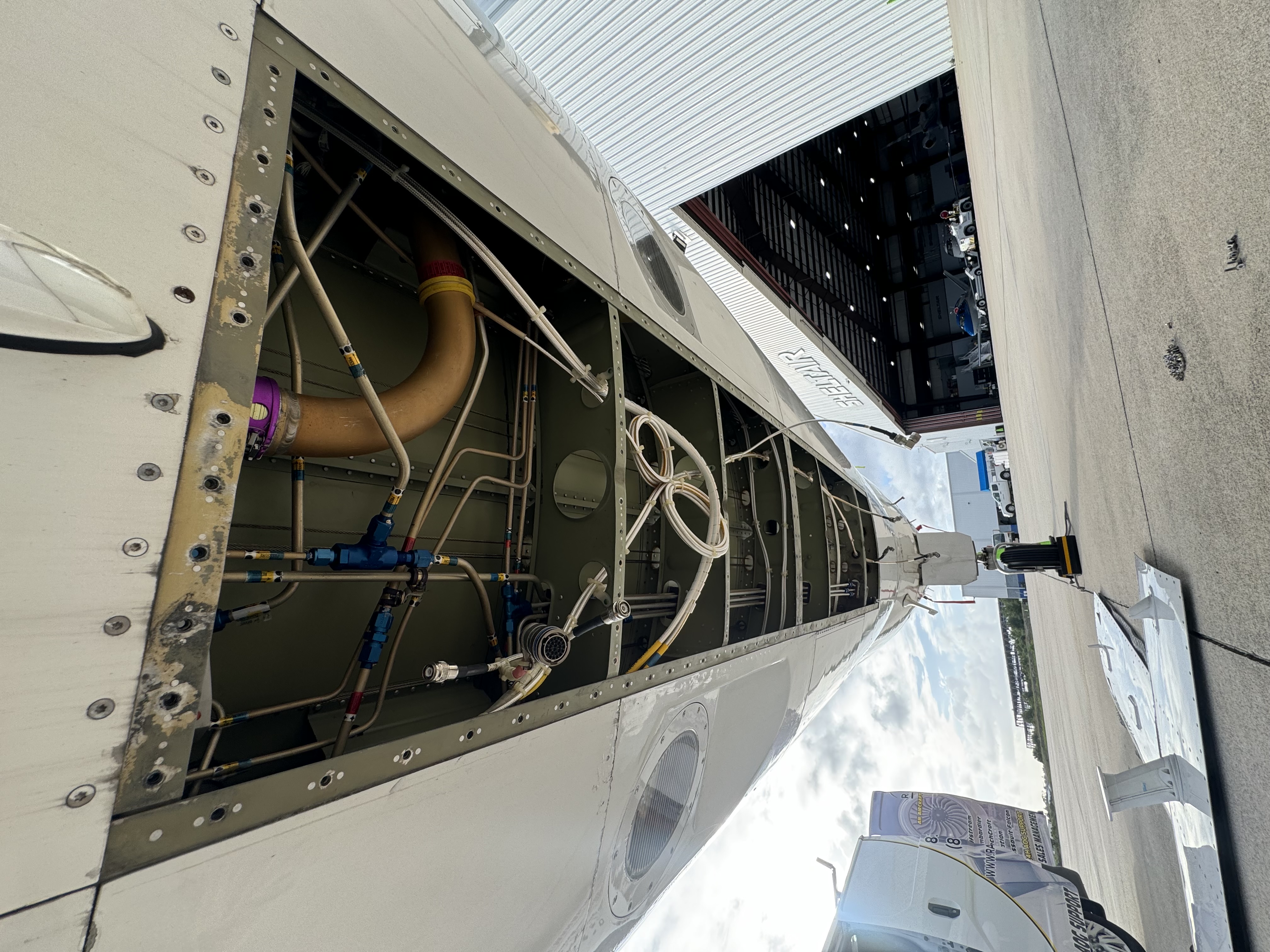 Underside view of an aircraft wing with the maintenance panel open, showing internal pipes, wires, and components near an airport hangar.