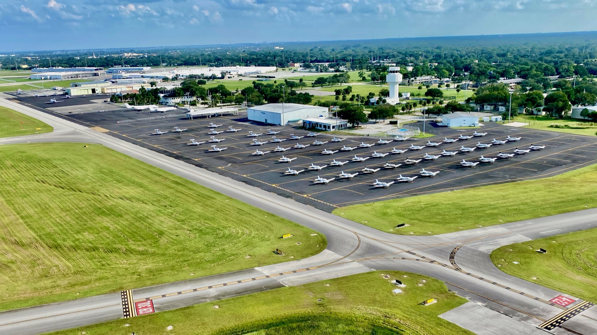 Aerial view of a small airport with multiple small airplanes parked in neat rows on the tarmac, surrounded by green grass and various buildings including a control tower.