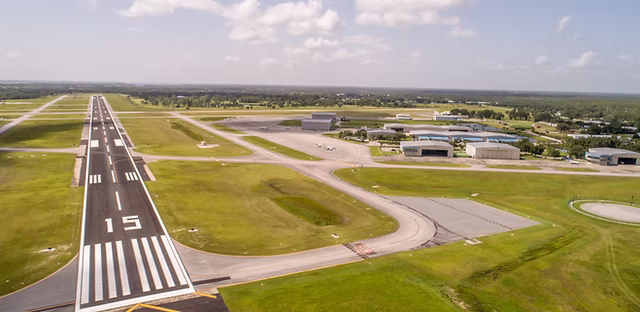 Aerial view of an airport runway labeled 15 with taxiways and several hangars in the background under a partly cloudy sky.