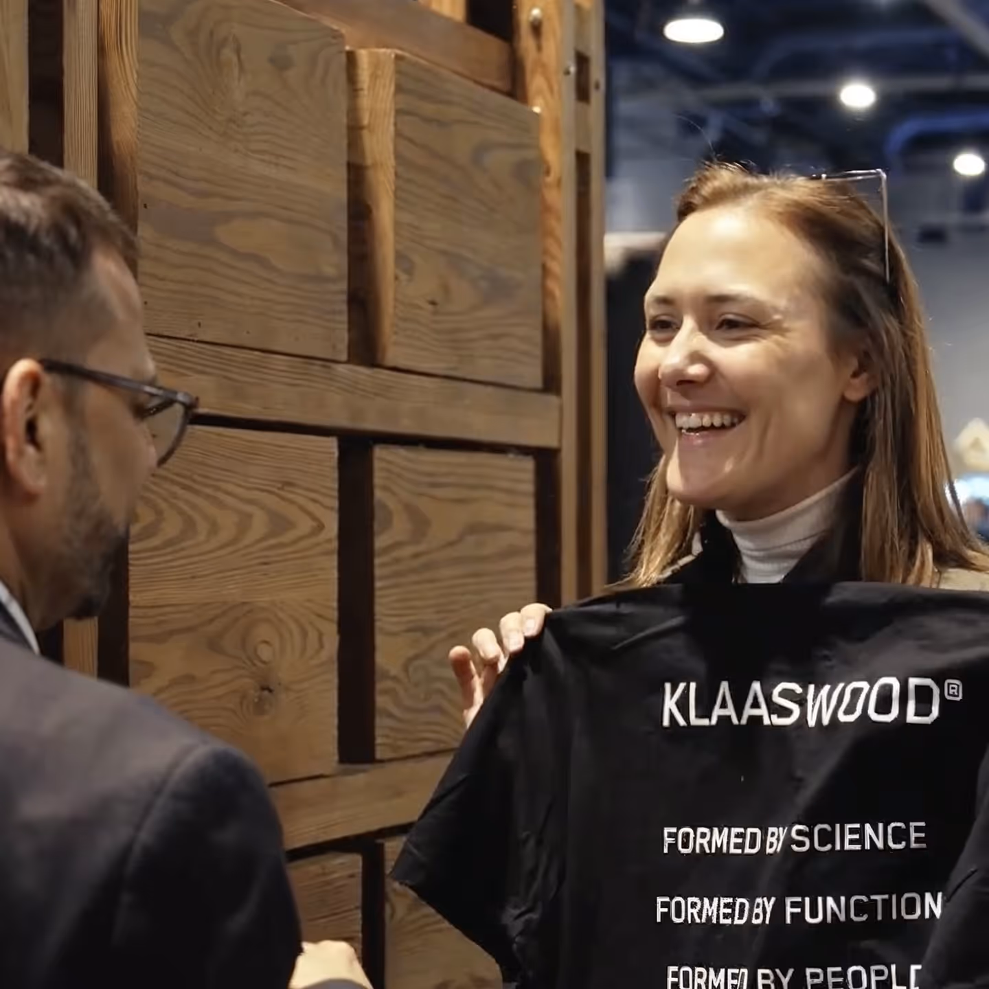 A smiling woman holds up a black T-shirt with white text that reads 'KLAASWOOD FORMED BY SCIENCE FORMED BY FUNCTION FORMED BY PEOPLE' while a man with glasses looks on.