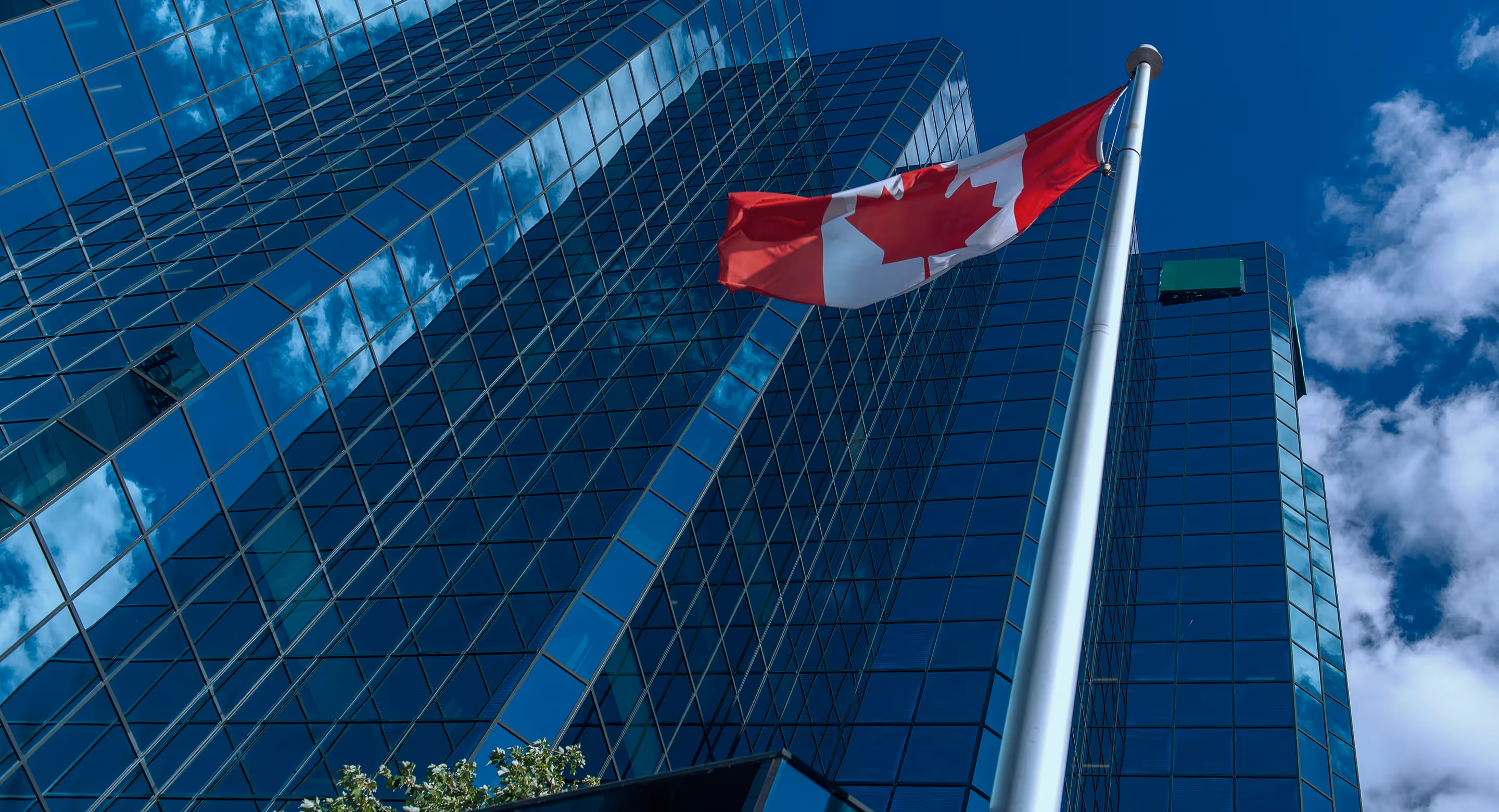 Canadian flag waving on a tall pole in front of a modern glass skyscraper reflecting the sky and clouds.
