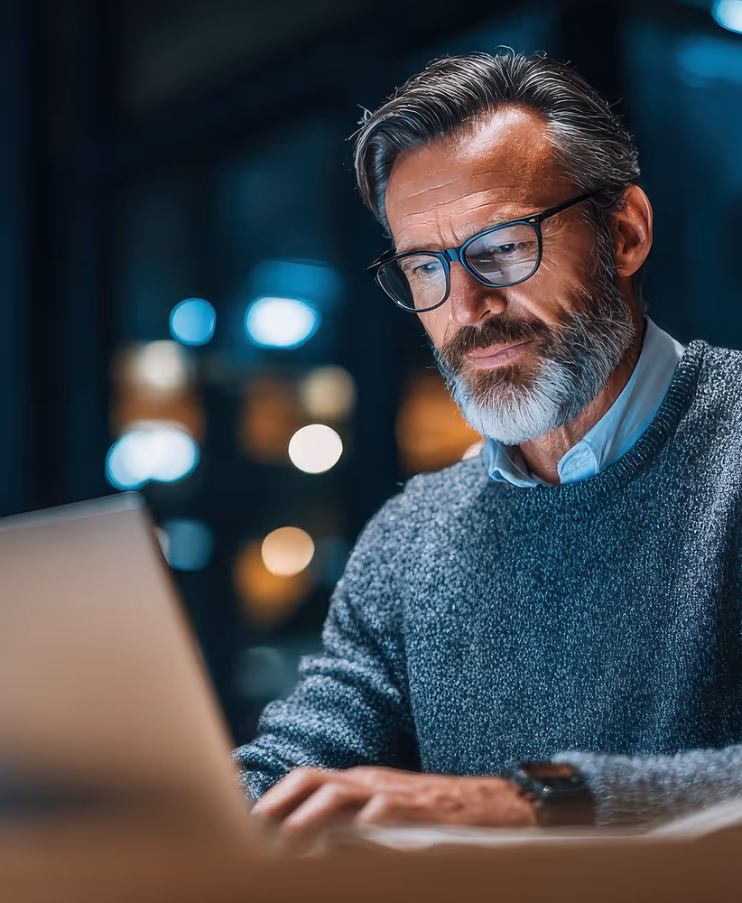Middle-aged man with glasses and gray beard working on a laptop in a dimly lit room.