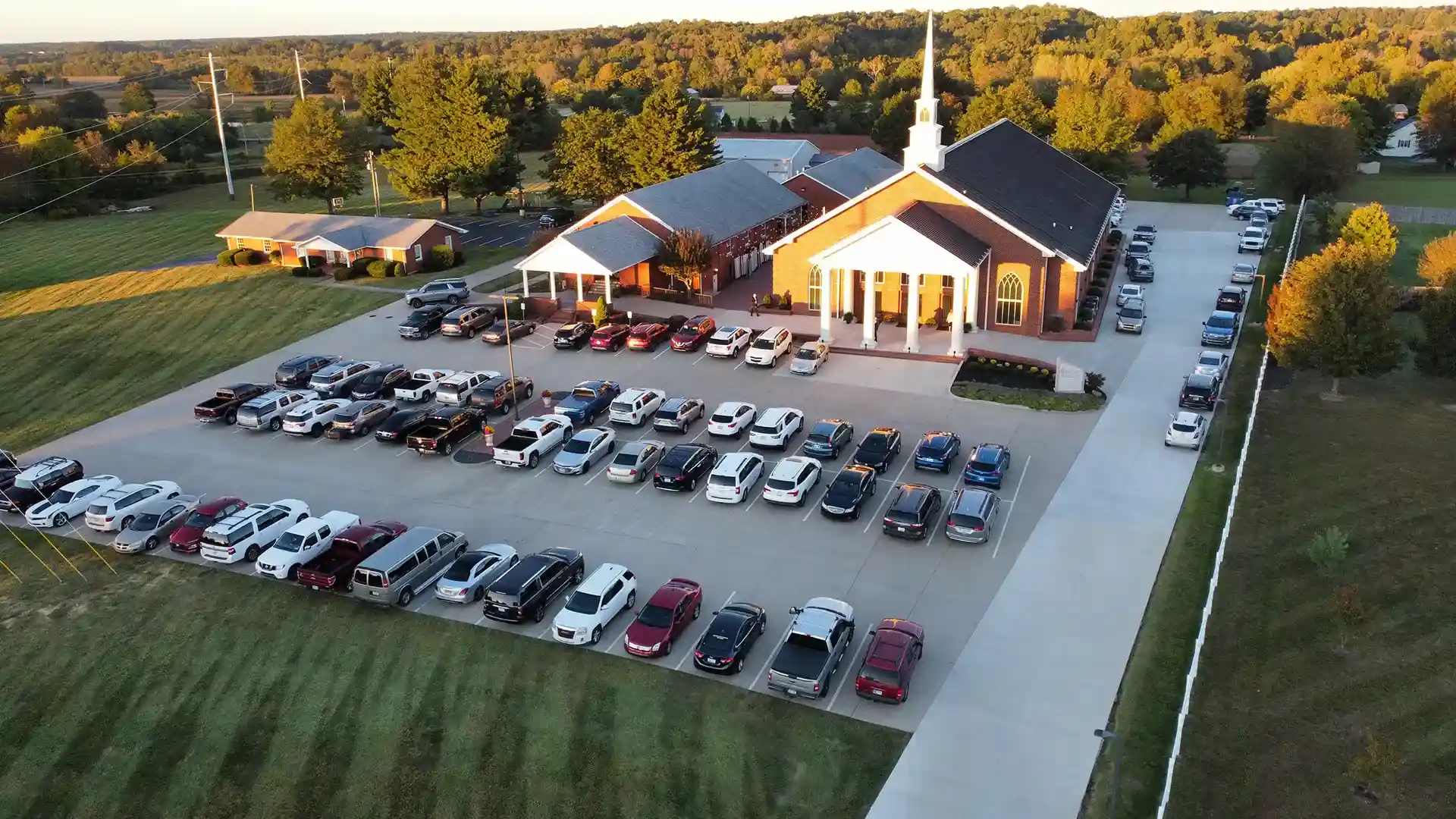 Aerial view of a church with a white steeple, surrounded by a large parking lot filled with cars and bordered by trees and fields.