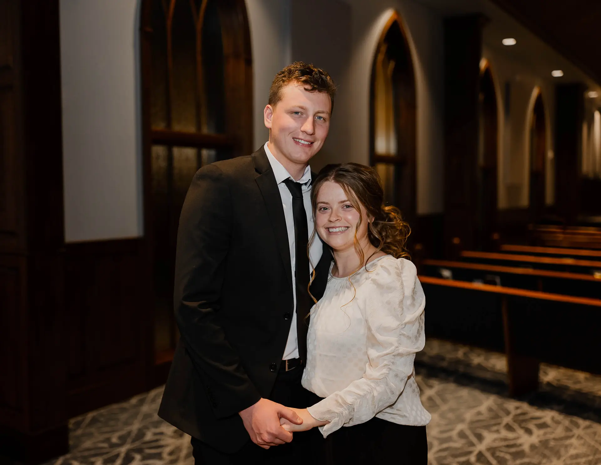 Smiling young man in a black suit and woman in a white blouse holding hands inside a church with wooden pews and stained glass windows.