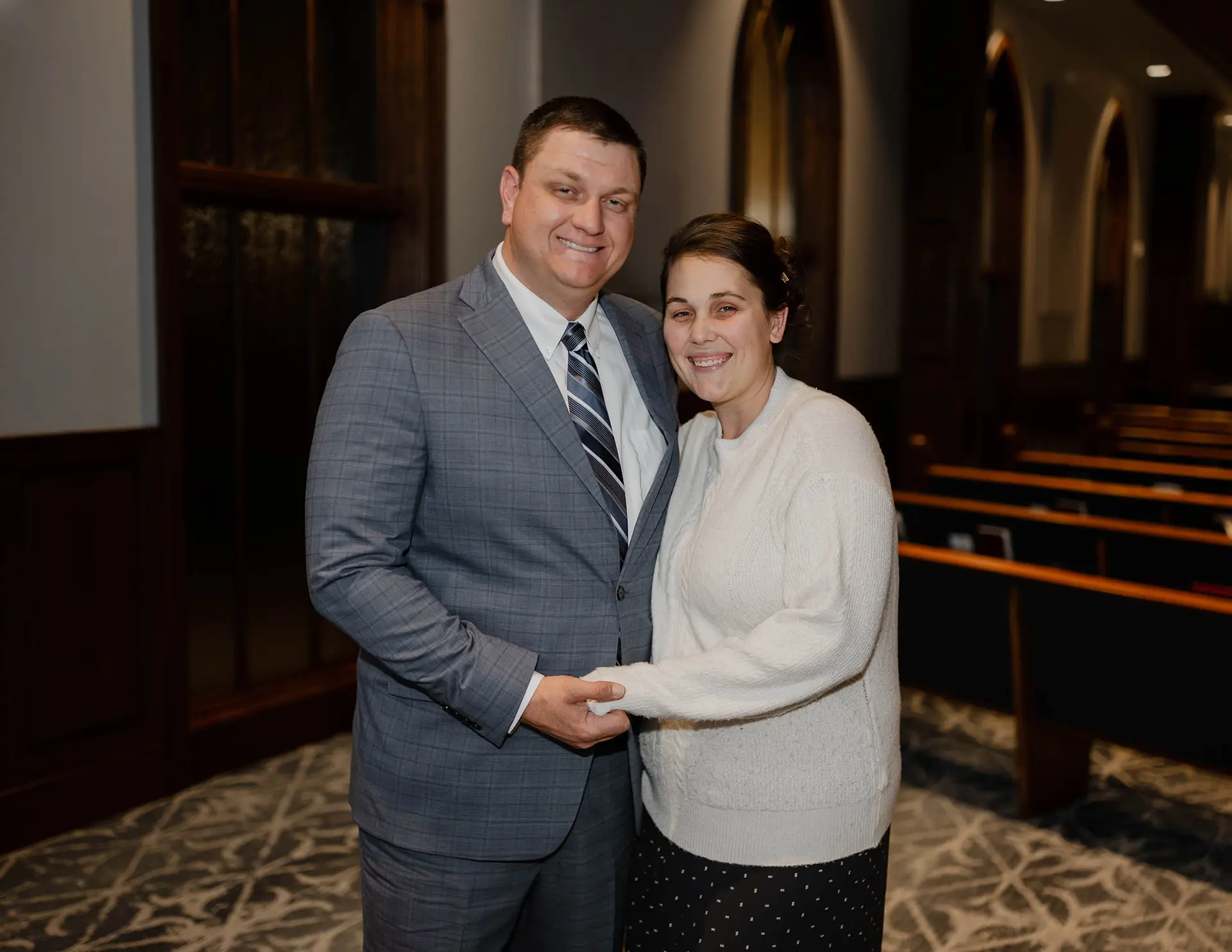 Man in a gray plaid suit and woman in a white sweater smiling and holding hands inside a church.