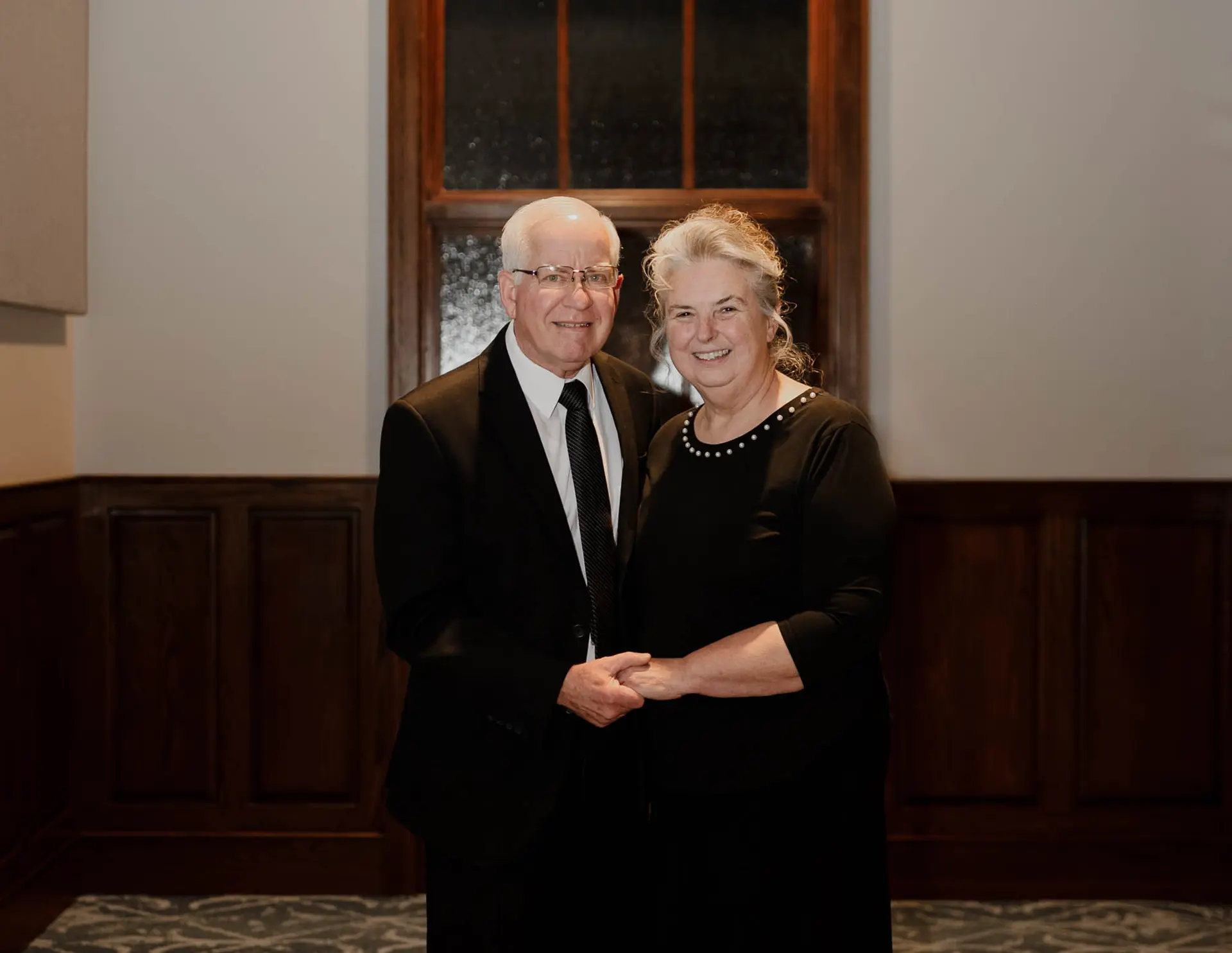 Elderly man in a black suit and tie holding hands with an elderly woman in a black dress, both smiling and standing indoors.