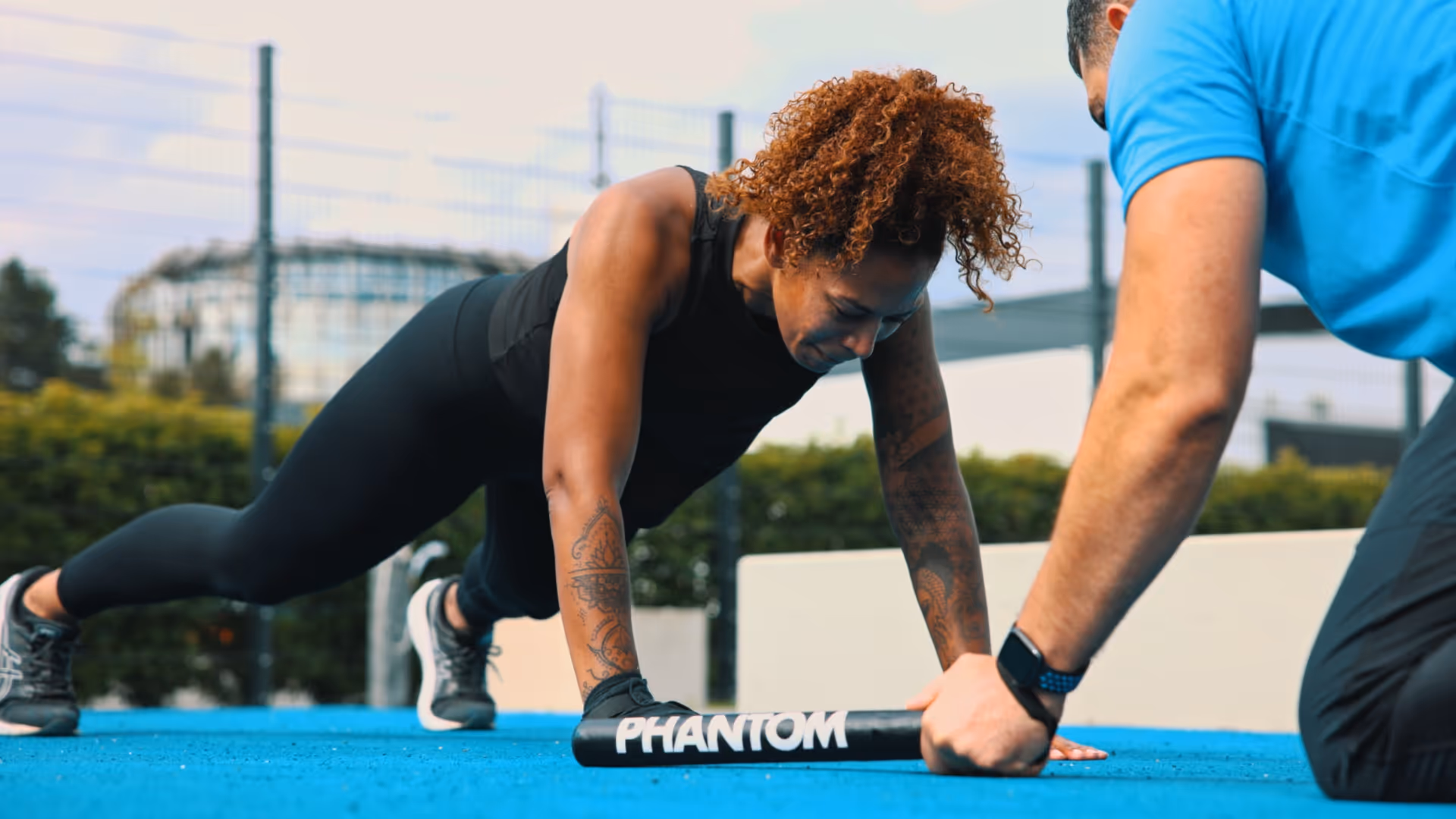 Frau macht Liegestütze auf blauem Boden, dabei hält ein Trainer eine Stange mit der Aufschrift ‚PHANTOM‘.