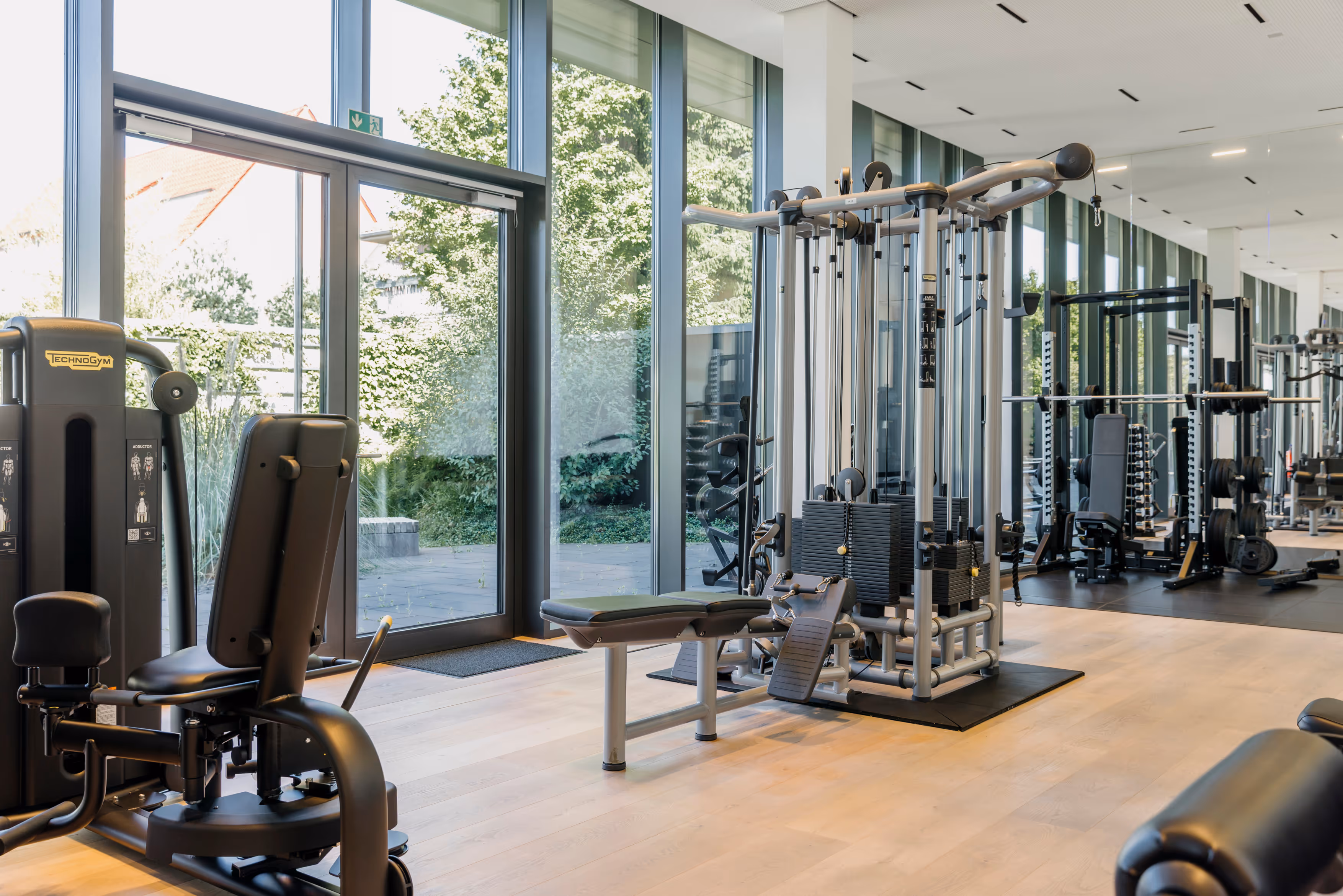 Helle und moderne Fitnessstudio mit Krafttrainingsgeräten und großen Fenstern mit Blick ins Grüne.