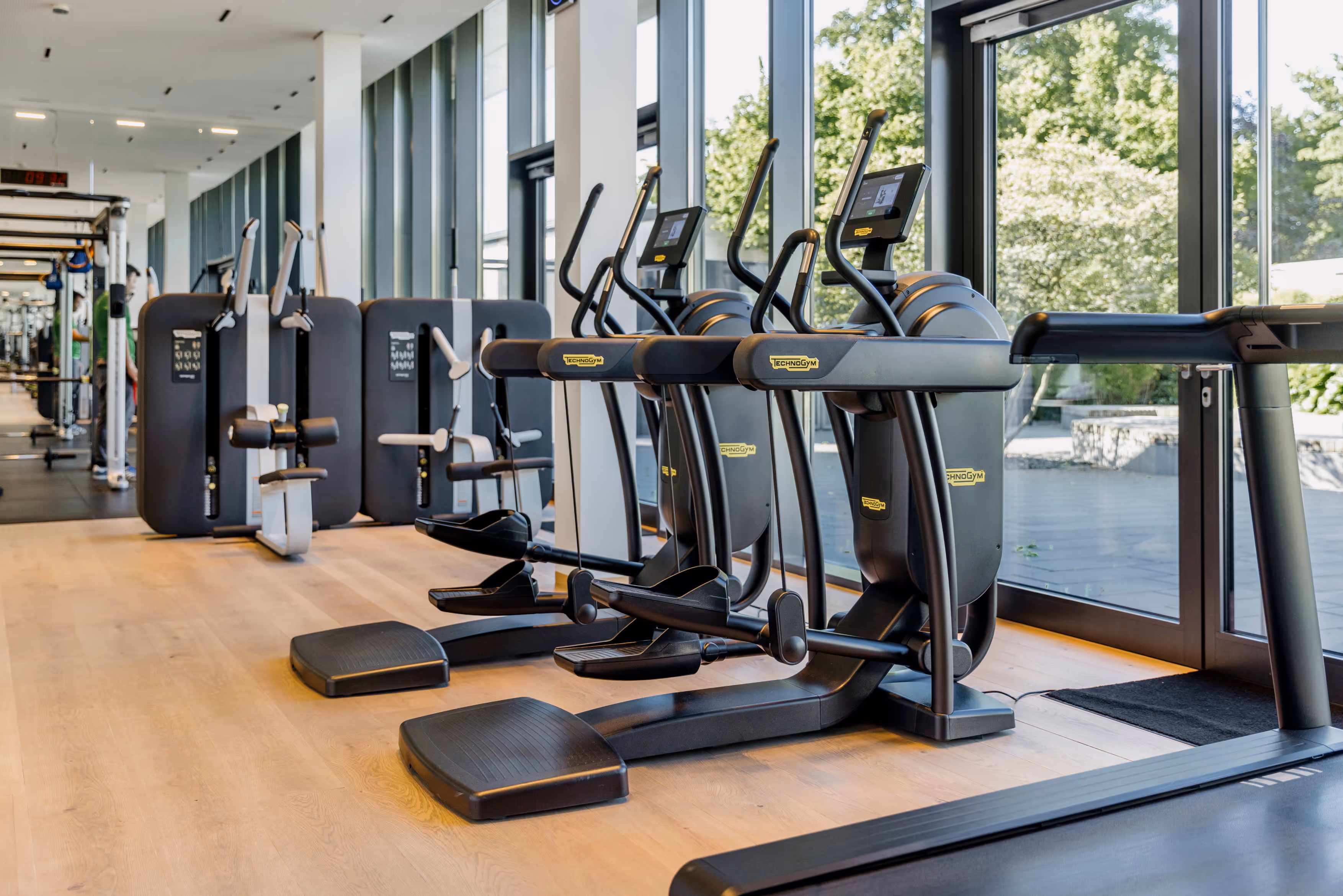 Moderne Fitnessgeräte in hellem Trainingsraum mit großen Fenstern und Blick auf grüne Bäume.