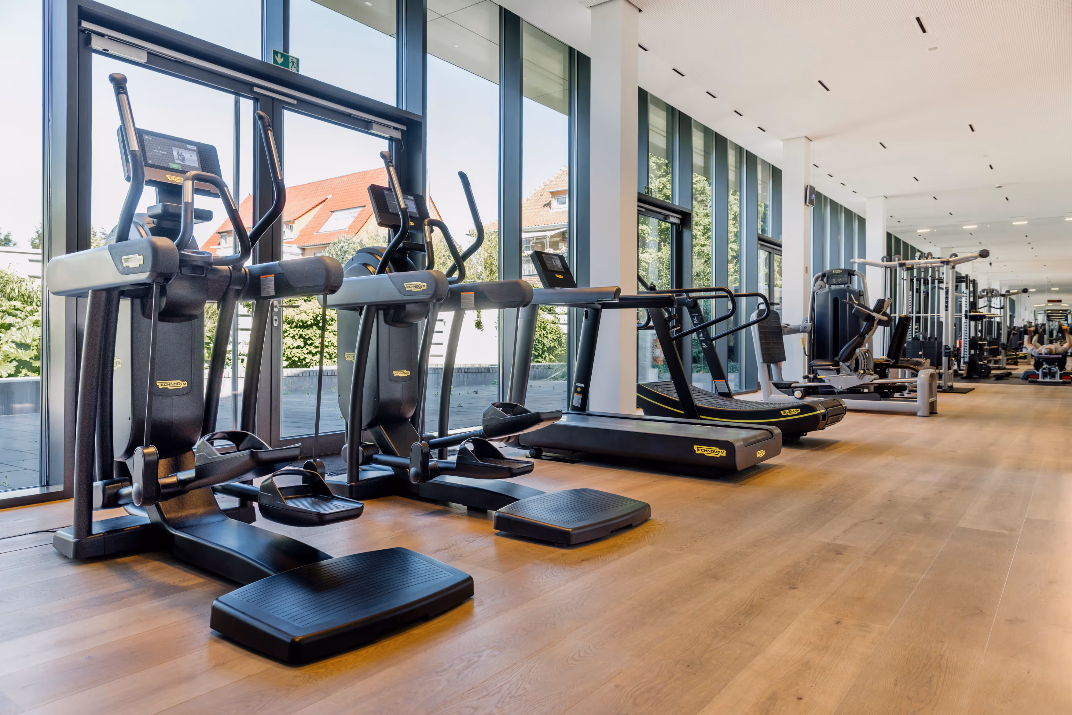 Modernes Fitnessstudio mit Trainingsgeräten und großen Fenstern, die Tageslicht hereinlassen.