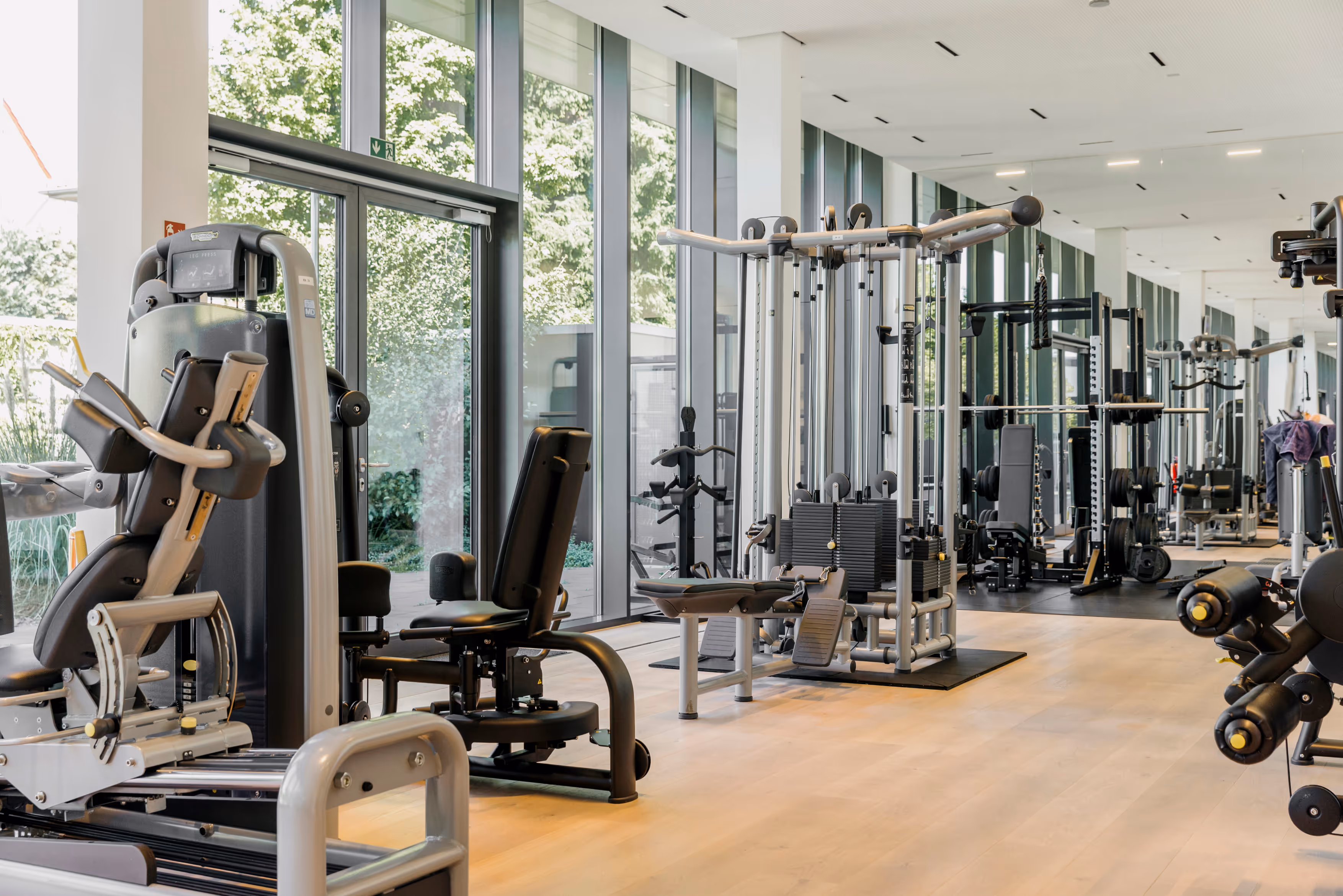 Heller Fitnessraum mit modernen Krafttrainingsgeräten und großen Fenstern mit Ausblick ins Grüne.