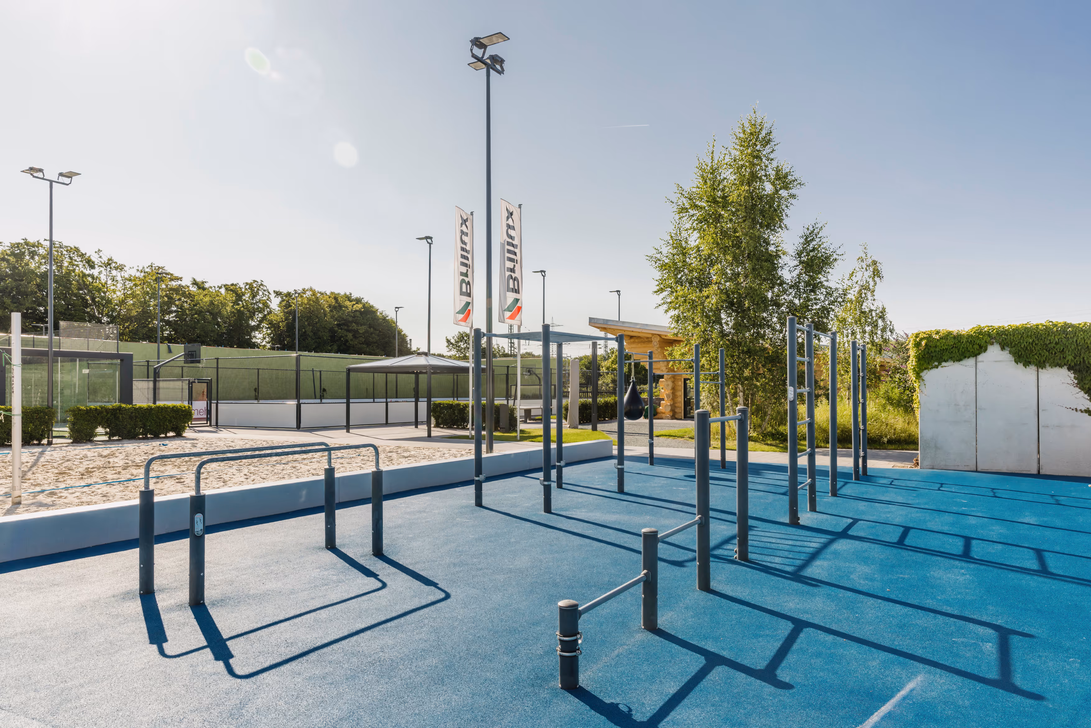 Moderner Fitnessparcours im Freien mit blauen Boden, Klimmzugstangen und Sandbereich unter klarem Himmel.
