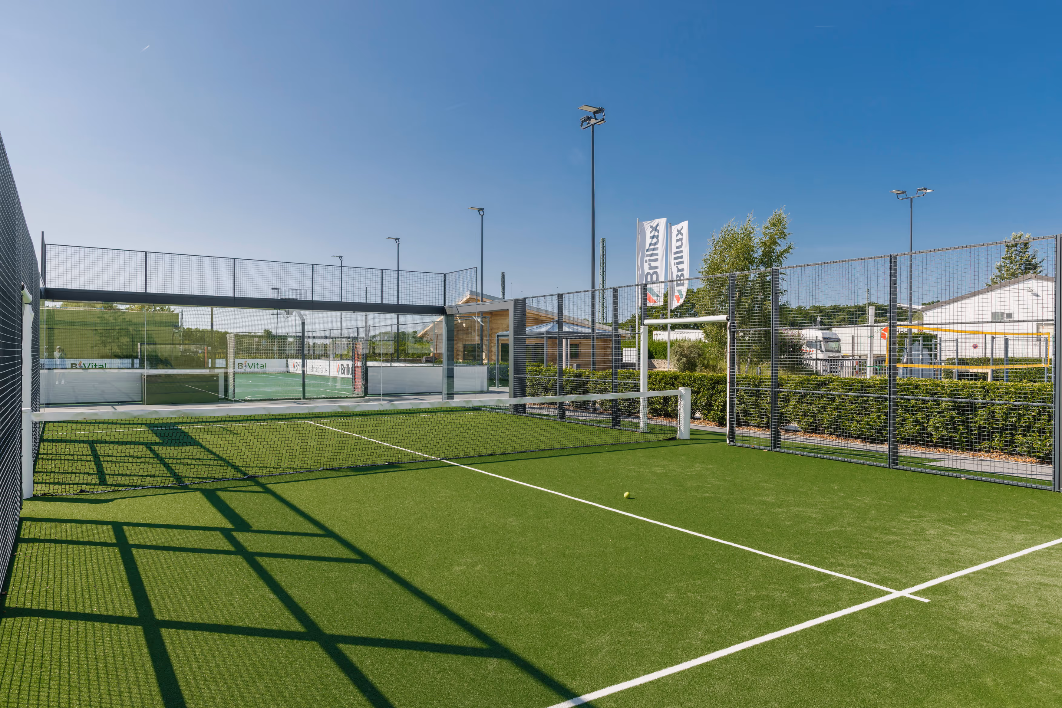 Tennisplatz mit grünem Kunstrasen, umzäunt von Metallgittern, einer Netzlinie und klarem blauem Himmel.