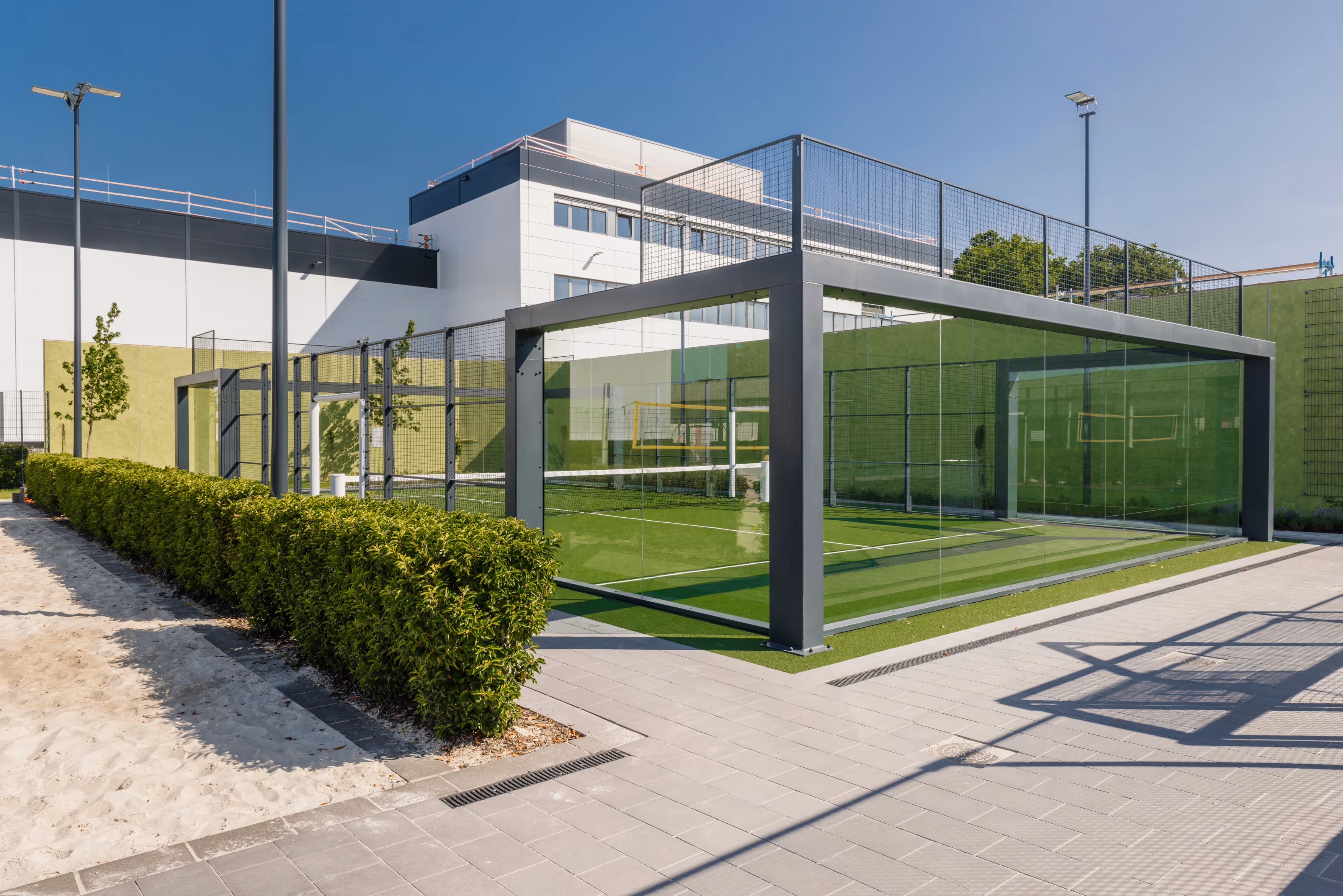 Moderner Padel-Tennisplatz mit grübem Kunstrasen, Glaswänden und sandigem Weg in einem Sportpark bei Sonnenschein.