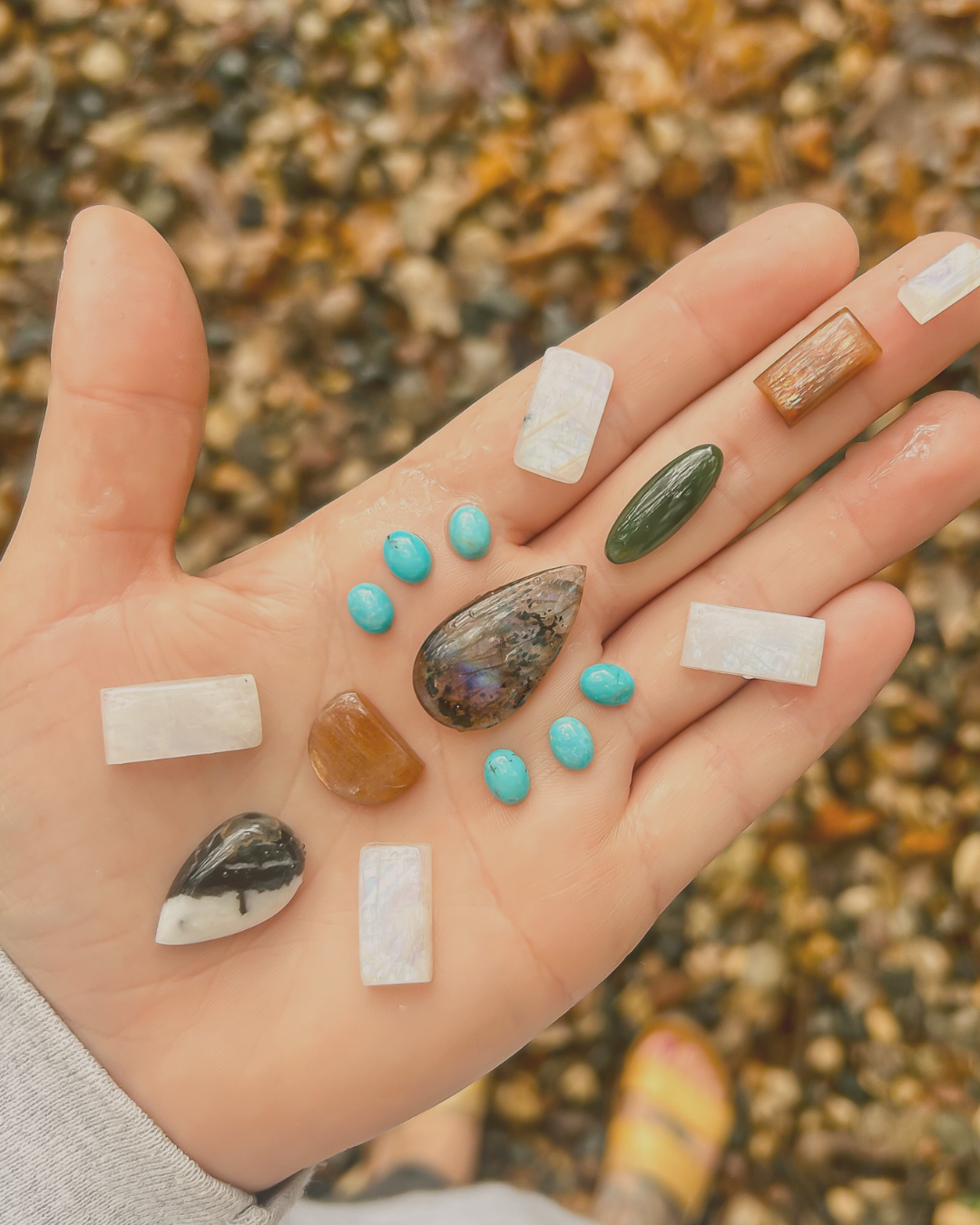 Open palm holding an assortment of polished stones including turquoise, moonstone, and jasper against a blurred pebble background.