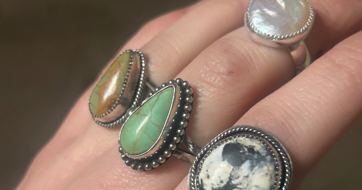 Close-up of a hand wearing four silver rings with different shaped stones including turquoise, jasper, mother of pearl, and a black and white stone.