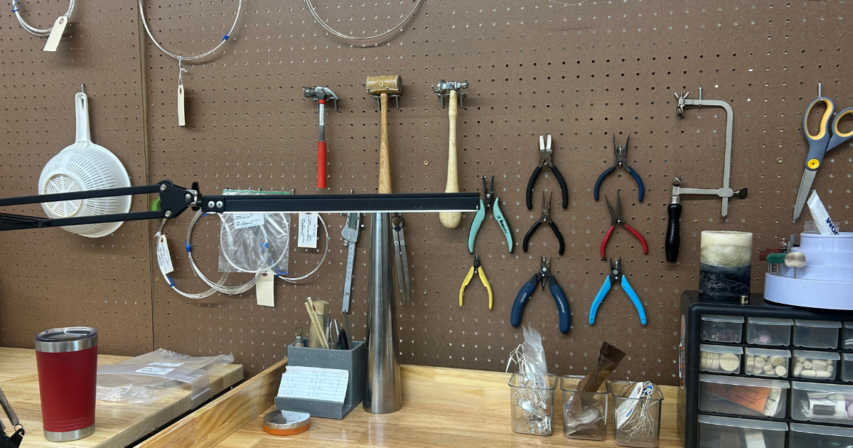 Workbench with hanging pliers, hammers, scissors, and wire on a brown pegboard, with various small tools and containers on the wooden surface.