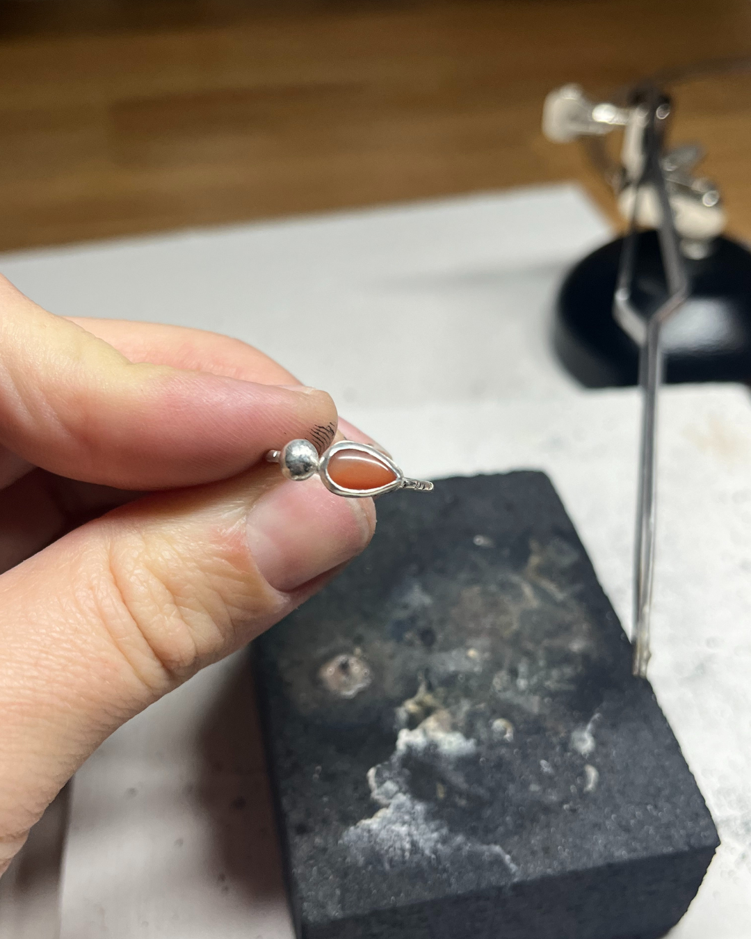 Hand holding a silver ring with a teardrop-shaped orange stone, above a black work surface and tool.