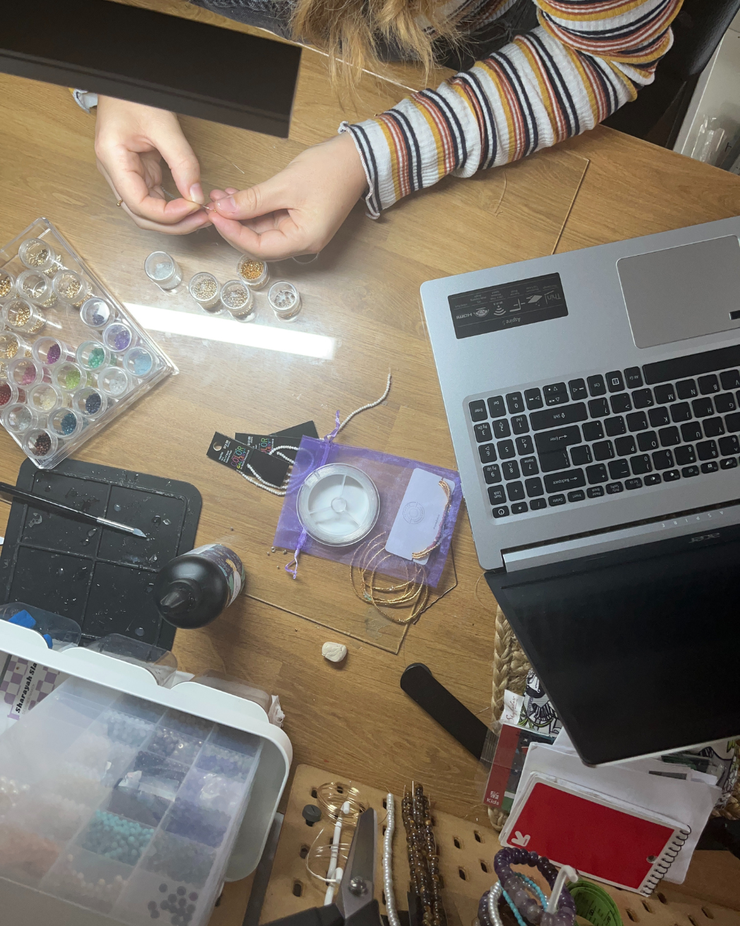 Person threading beads at a wooden desk with a laptop, containers of beads, string, and jewelry-making tools.