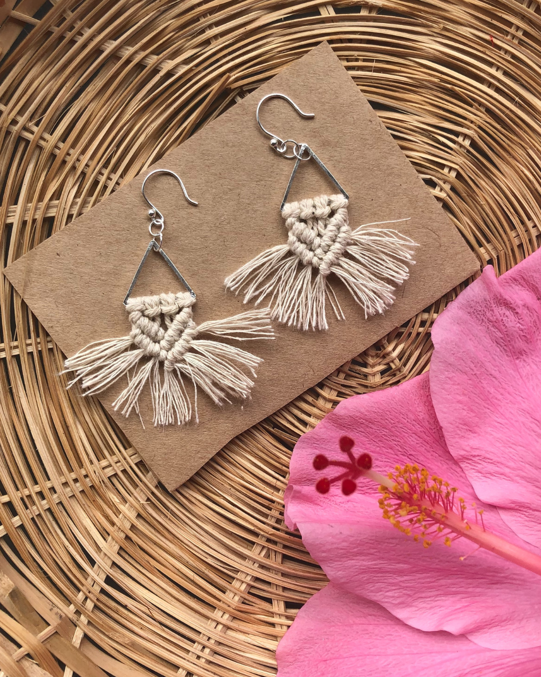 Handmade macrame earrings with silver hooks displayed on brown paper over a woven rattan surface, next to a pink hibiscus flower.
