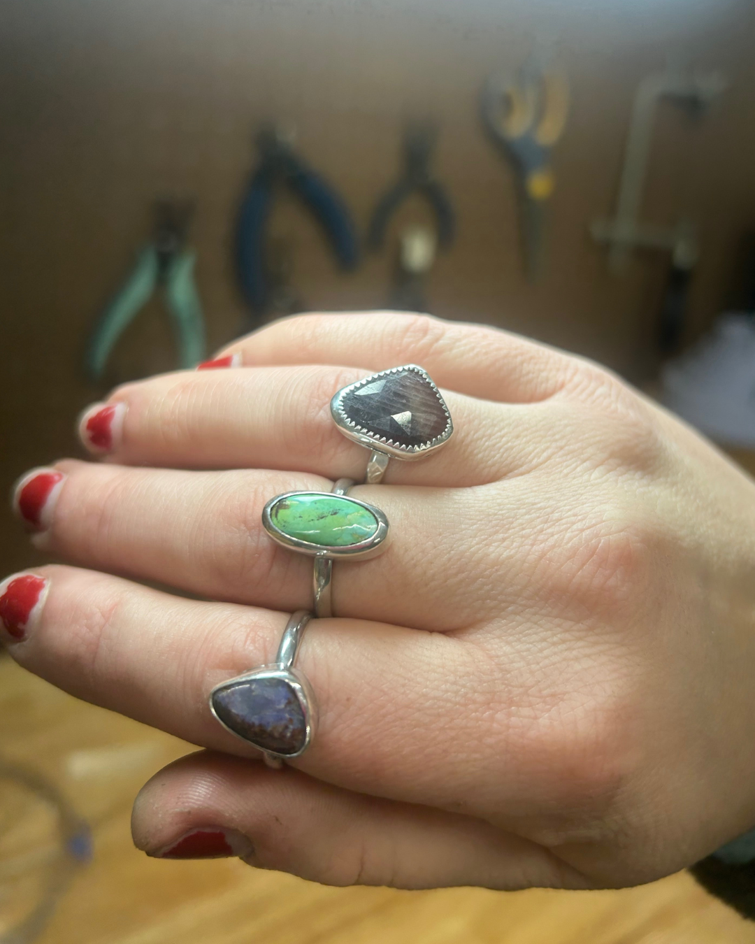 Hand with red nail polish wearing three silver rings, each with a differently colored stone: one purple, one green, and one dark gray.