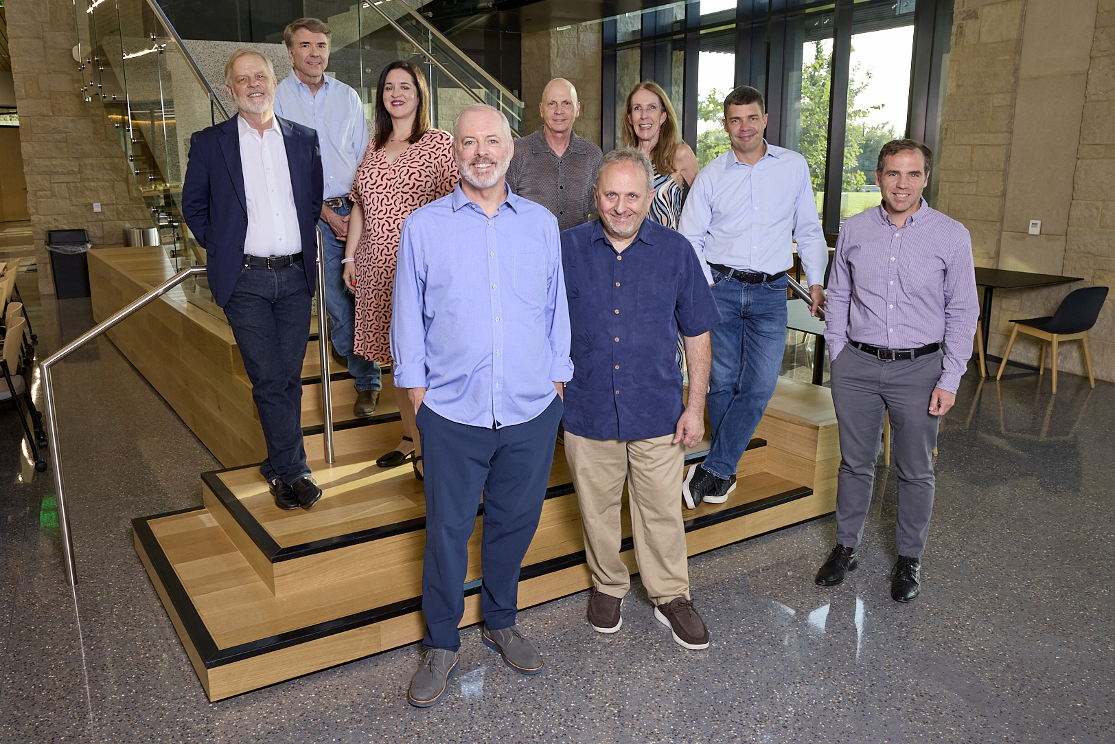 Group of eight men and two women standing on and around wooden stairs inside a modern building with large windows and stone walls.