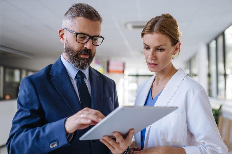 Man in a suit and glasses showing a tablet to a female healthcare professional in a white coat.