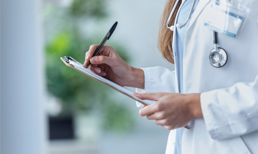 Close-up of a healthcare professional in a white coat holding a pen and writing on a clipboard.